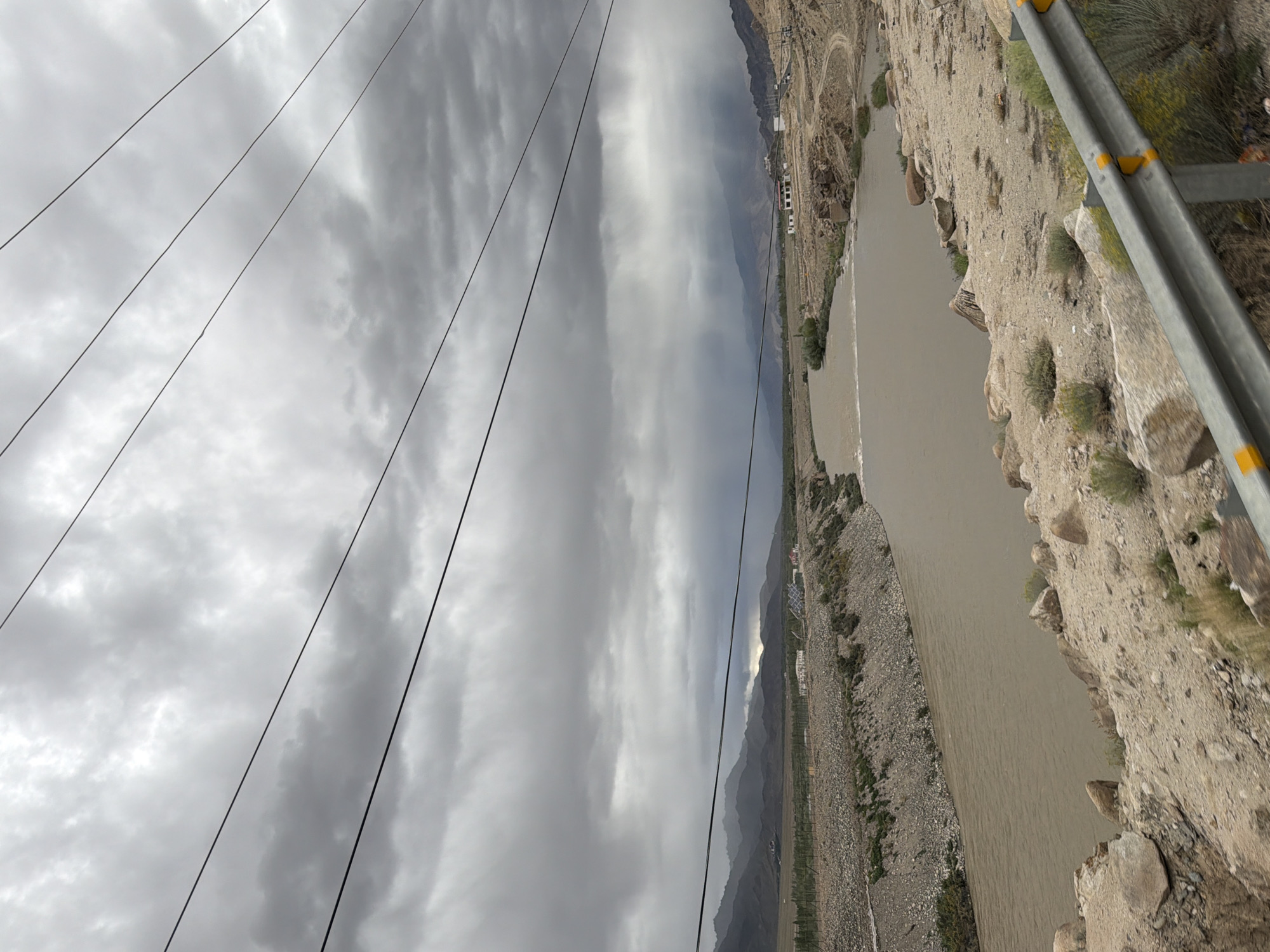 The image depicts a wide, muddy river flowing through a dry, mountainous landscape. The sky is overcast with heavy, gray clouds, suggesting impending or ongoing rain. Power lines are visible in the foreground, crossing the frame horizontally. The riverbanks are lined with rocky terrain and sparse vegetation. In the distance, rolling hills and mountains form the backdrop, with a few scattered buildings visible on the plains. The overall scene conveys a sense of remoteness and a rugged natural environment.