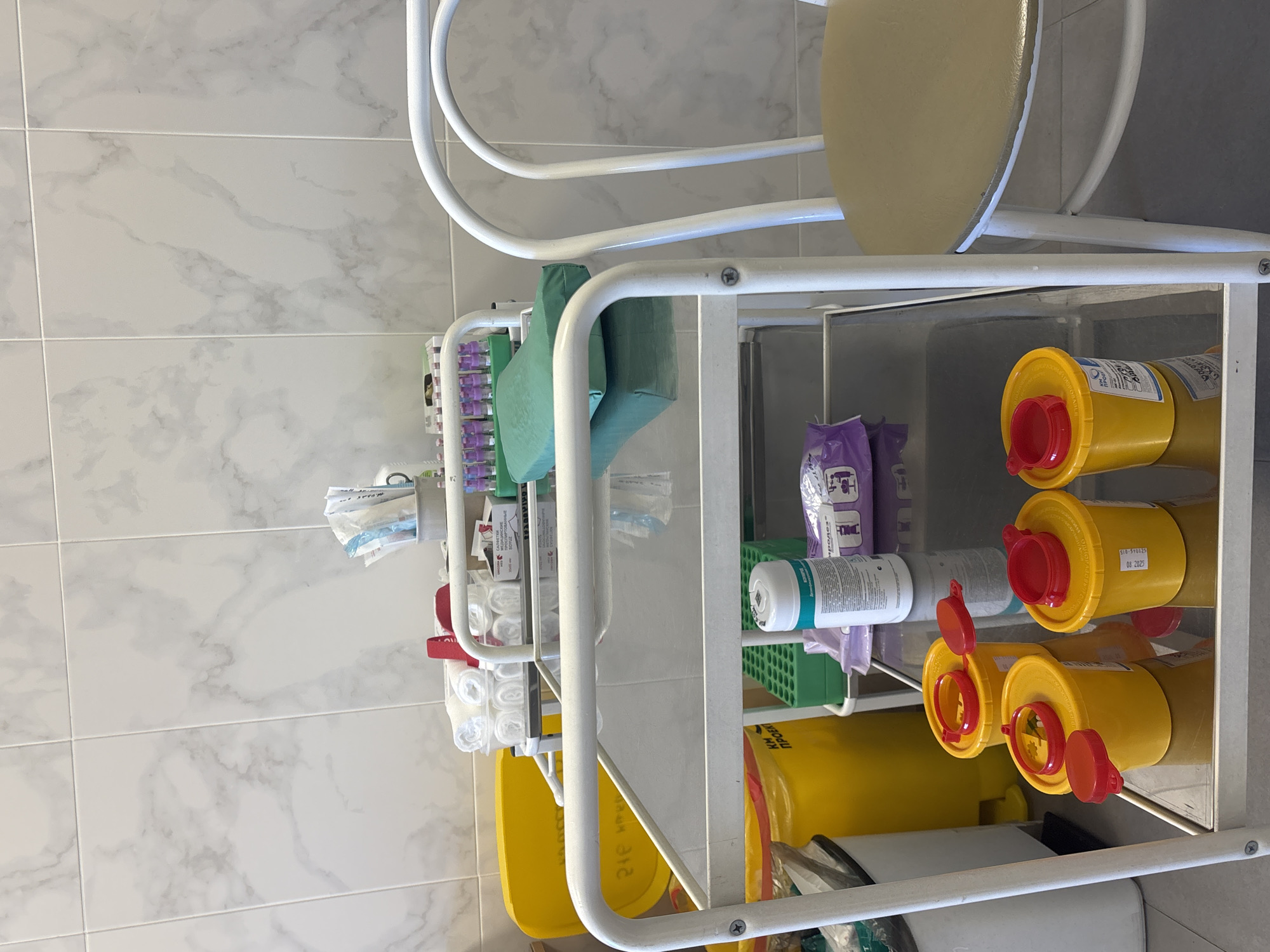 The image depicts a medical cart in what appears to be a doctor's office or clinic. The cart is white and has multiple shelves holding various medical supplies. Some of the items on the cart include:
* Vacutainer tubes and tourniquets.
* Sterile supplies, possibly bandages.
* Sharps containers, which are yellow with red lids.
* A container of disinfectant wipes.
* Other medical consumables.
The presence of Russian text on some of the labels suggests the location is in Russia. Given the request for Saint Petersburg, it's plausible the image was taken in a medical facility there.
The environment appears clean and well-maintained, typical of a healthcare setting. The background shows tiled walls, suggesting a clinical or laboratory environment.