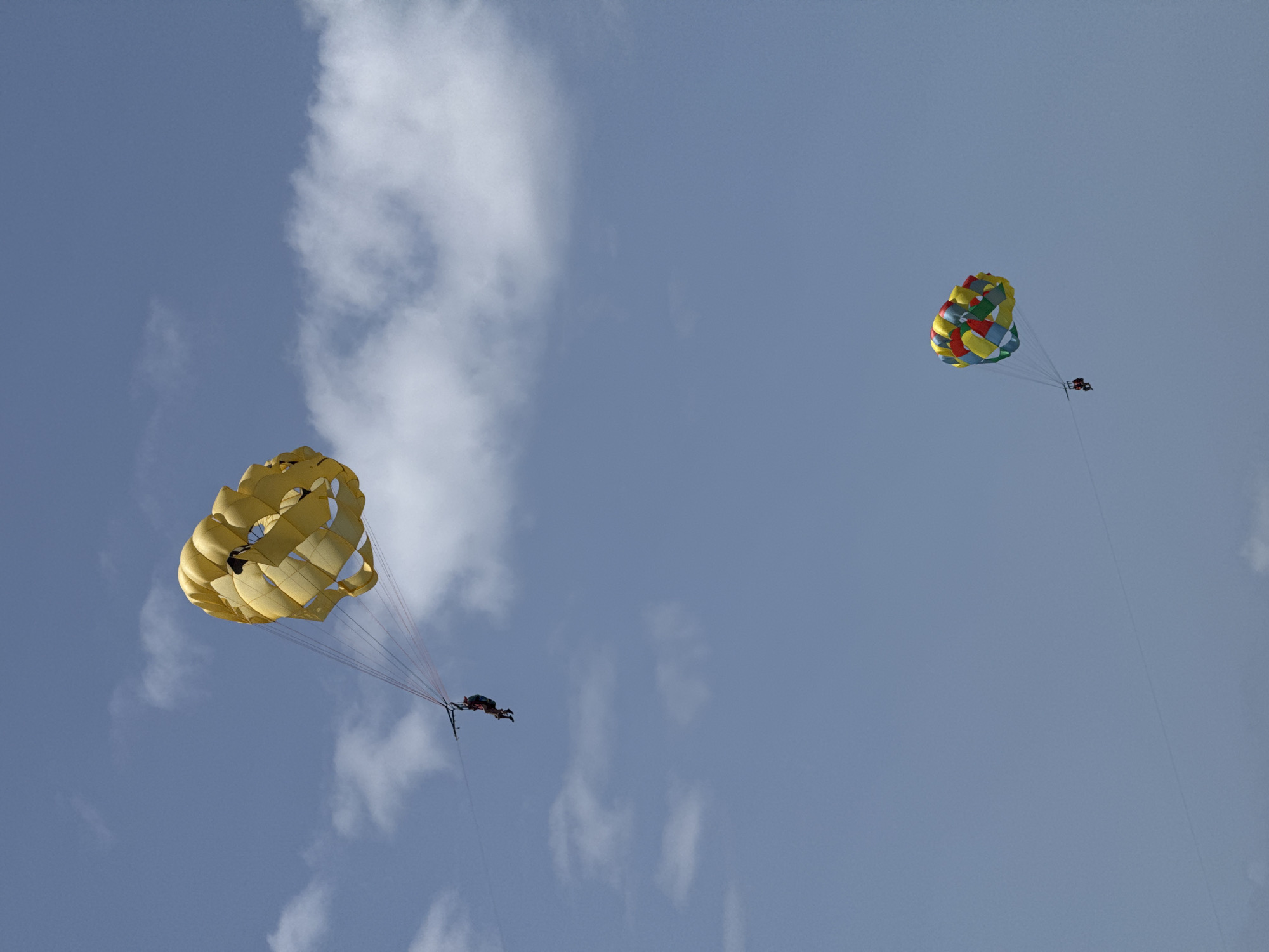 Two parascenders are seen against a bright blue sky with scattered white clouds. The parascenders are attached to colorful parachutes that catch the wind, lifting them high above. One parachute, predominantly yellow, is closer to the foreground and appears larger, with its fabric billowing. The other parachute, smaller and further in the distance, is a vibrant mix of red, yellow, green, and blue, creating a patchwork effect.
Each parachute is tethered to a person or persons who are enjoying the exhilarating experience of parasailing. They appear to be suspended in the air, possibly being towed by a boat out of frame. The scene suggests a sunny day, typical for a tropical location like Higüey, Dominican Republic, where this activity is a popular tourist attraction. The clear sky and good visibility indicate favorable weather conditions for such an adventure. The overall mood is one of freedom and excitement, as the individuals soar through the air, taking in the expansive views. No visible text is present in the image.