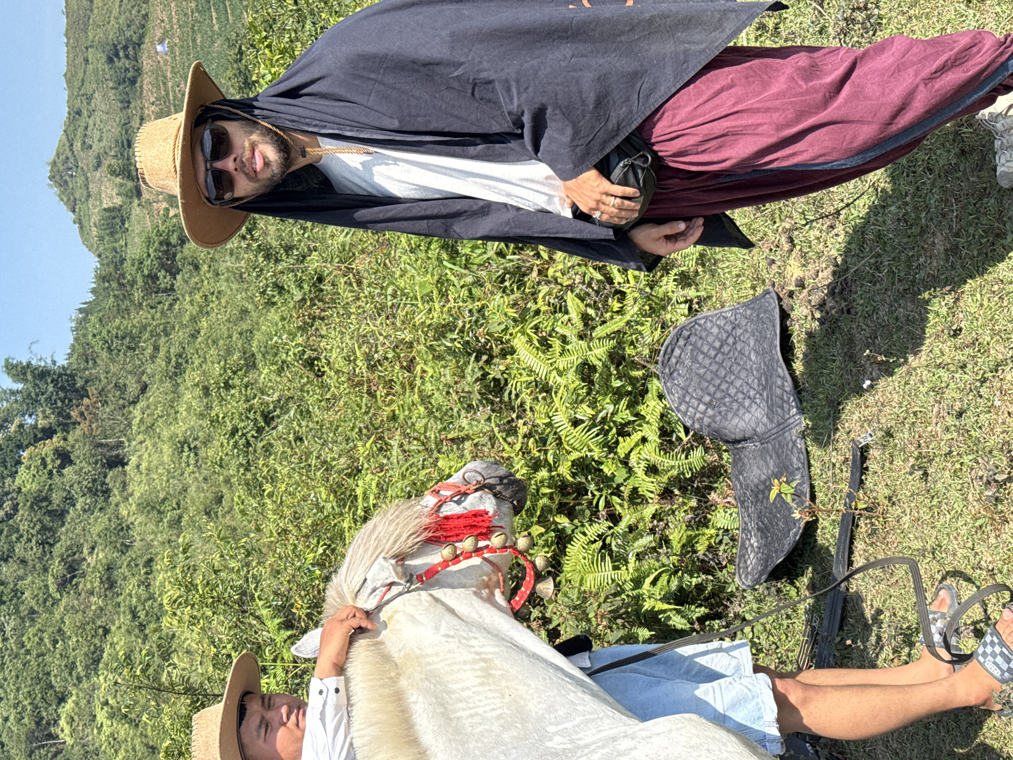 In a medium shot taken outdoors in Bắc Hà, Vietnam, two individuals and a white horse are the subjects of the image.
One person, wearing a straw hat and sunglasses, is draped in a dark cloak over a white shirt and maroon trousers. A black pouch hangs from the person's waist. This person is lying down on the grass.
The second person is also wearing a straw hat. This person is seated in a light blue garment and a white shirt. This person is holding a white horse with a decorative red bridle adorned with small bells.
A black saddle lies on the ground between the individuals and the horse. The environment consists of green vegetation, suggesting a field or rural area in Bắc Hà, Vietnam. Hills are visible in the background.