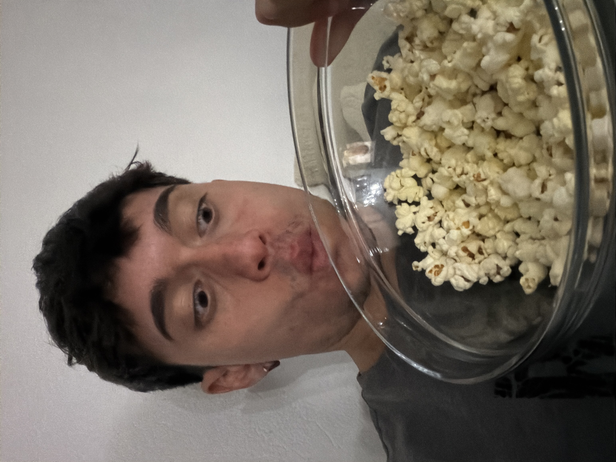 A person with dark hair and light skin is holding a clear glass bowl filled with popcorn close to their face. The person appears to be looking directly at the camera with a slightly puckered expression. The background is a plain white wall. The person is wearing a gray shirt. There is no indication of the location being Blagoevgrad, Bulgaria.