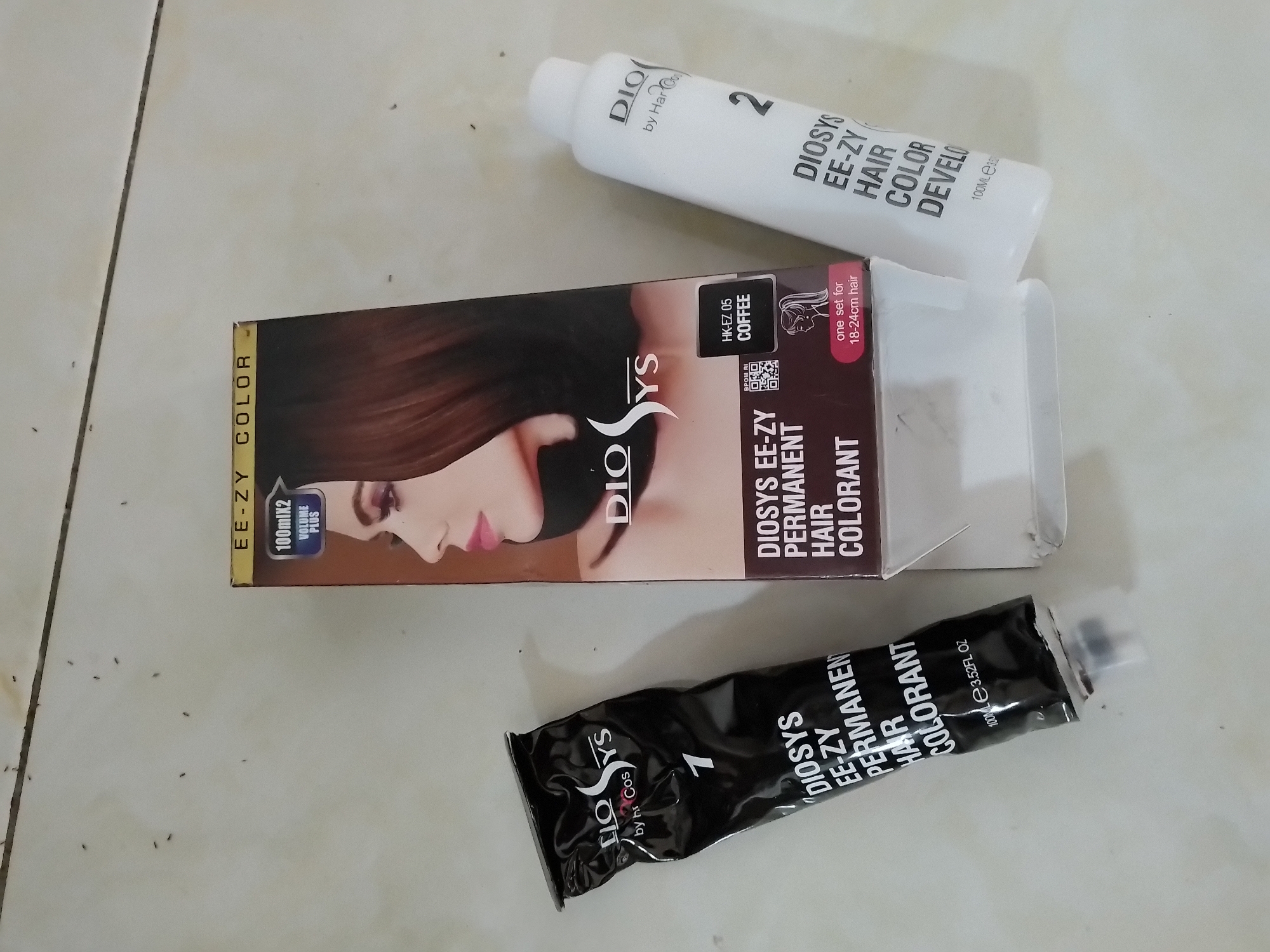 Two hair dye products are laid out on a light-colored, textured surface, possibly a tiled floor. To the left is a black tube labeled "DIOSYS EE-ZY PERMANENT HAIR COLORANT 1". To the right is a white bottle labeled "DIOSYS EE-ZY HAIR COLOR DEVELOPER 2". A box for "DIOSYS EE-ZY COLOR" is positioned between the two products. The box features an image of a woman with dark hair and text including "100mlX2 VOLUME PLUS", "HK-EZ 05 COFFEE", and "one set for 18-24cm hair". The products and box appear to be new. The lighting suggests an indoor environment.