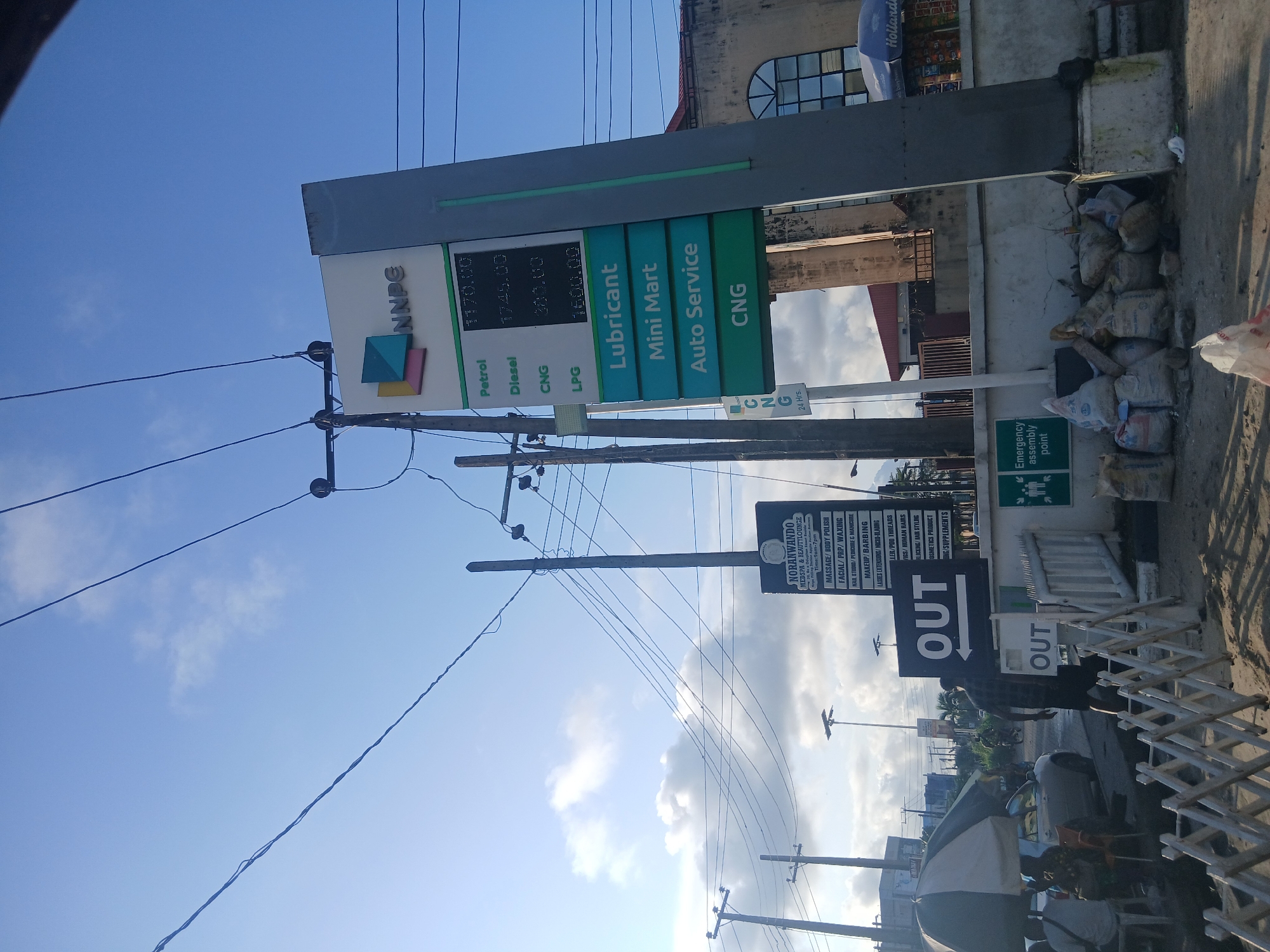 The image depicts a street scene in Lagos, Nigeria, likely a commercial area with a gas station. A prominent NNPC (Nigerian National Petroleum Corporation) gas station sign stands tall, displaying fuel prices for Petrol, Diesel, CNG, and LPG. Adjacent to the fuel price display, there are signs for Lubricant, Mini Mart, Auto Service, and CNG. To the left, a sign for "NORANWANDO FREDSPA & BEAUTILOUNGE" lists various beauty services offered. A large black sign with the word "OUT" and an arrow is also visible.
Several utility poles with extensive electrical wiring dominate the sky. The environment appears somewhat dusty with piles of sacks, likely refuse or construction materials, at the base of a wall. In the foreground, a white foldable barrier is set up. People are partially visible near a shaded area, with a car parked behind them. Further in the background, more vehicles and people can be seen, suggesting ongoing activity. The sky is blue with scattered clouds.