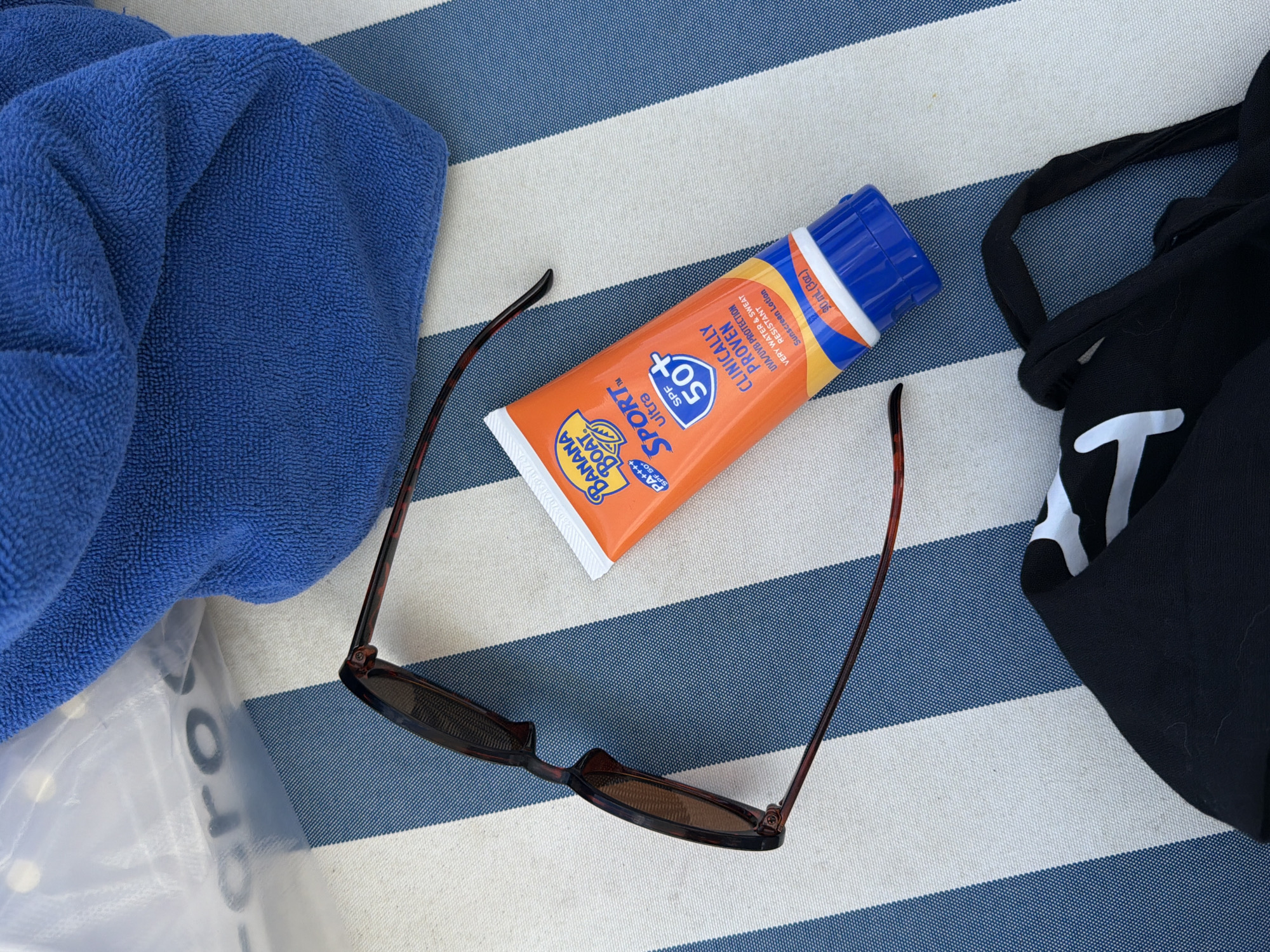 The image shows a still life composition of personal beach items arranged on a blue and white striped surface, possibly a beach towel or chair. On the left, a folded blue towel partially covers a clear plastic bag with a visible logo or text "Cro." Next to the towel lies a tube of Banana Boat Sport Ultra SPF 50+ sunscreen. A pair of brown sunglasses are positioned around the sunscreen. To the right, part of a black tote bag with a large white "T" is visible. The scene suggests a beach or poolside setting. There is no evidence to link it to Đà Nẵng, Vietnam.