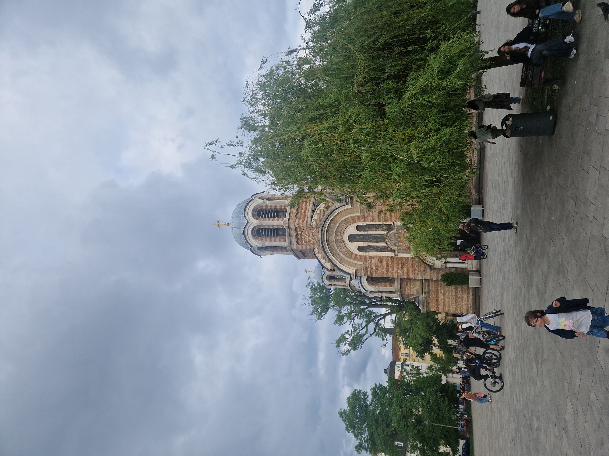 The image shows a tall Eastern Orthodox church with a domed roof on a cloudy day. The church has a brick facade and arched windows. A large weeping willow tree partially obscures the view of the church. On the right side of the image, a paved area features people relaxing on benches and walking by. Several children are seen on bicycles in the foreground. The location appears to be an urban public square.