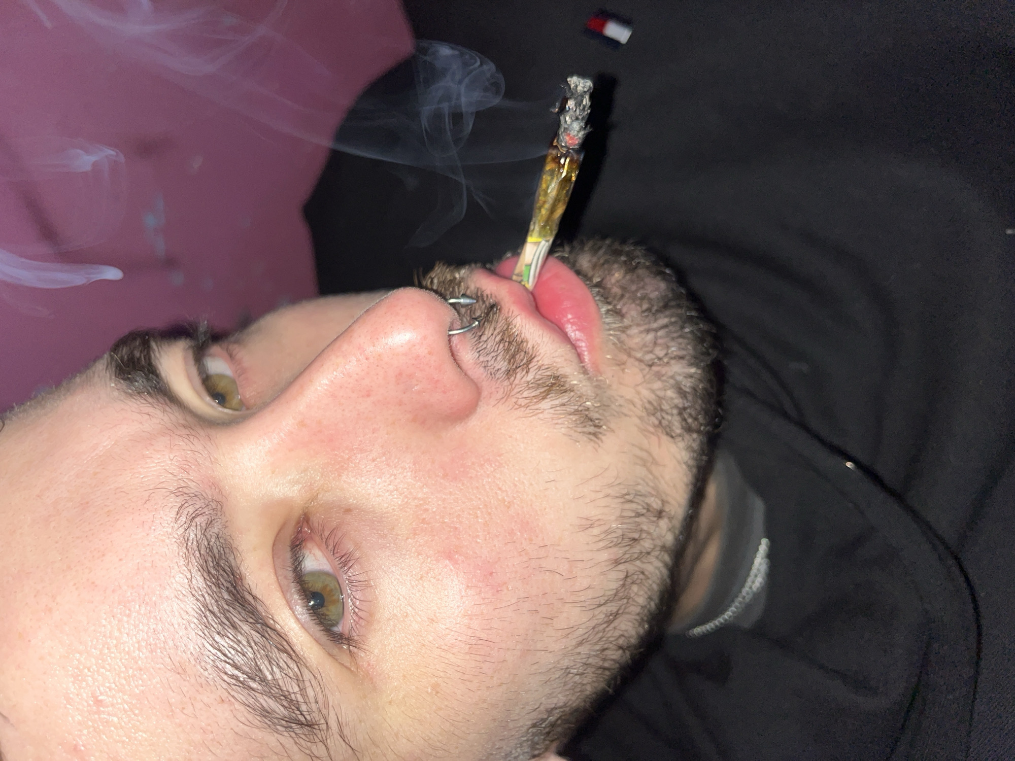 A close-up, tilted shot shows a person with light skin, stubble, and green eyes holding a lit joint in their mouth. The person has a silver hoop nose piercing. Smoke rises from the joint. The background is partially pink and partially dark, with what appears to be a Tommy Hilfiger logo visible near the dark area. The location is not discernible from the image provided.
