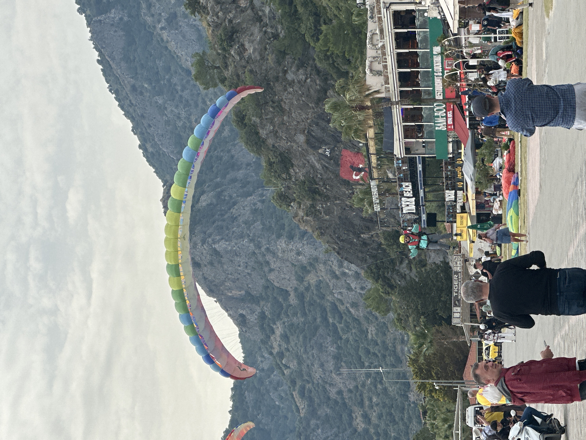 The image shows a scene in Fethiye, Turkey, recognizable by the Turkish flag. A paraglider with a multicolored parachute (yellow, green, blue, and red) is in the sky. In the background is a mountain. In the foreground, there is a crowded area with buildings. The buildings are lined with signs for various restaurants and bars, including "TIGER BEACH", "TIGER RESTAURANT & BAR", "Amarillo 66", "BAM-BOO RESTAURANT & LOUNGE", and "ROVAL ROCK". People are standing and walking around. Some are looking up at the paraglider. Some are wearing casual clothing. One person is wearing a red jacket. Another person is wearing a blue plaid shirt. The ground appears to be paved.