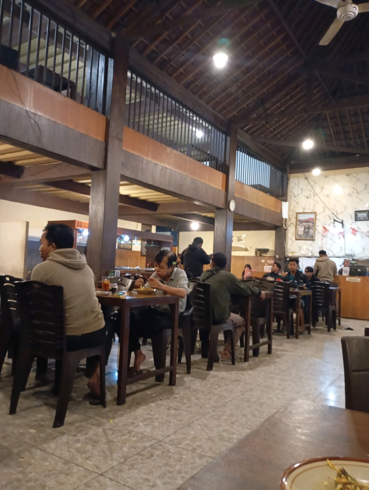 The image depicts the interior of a restaurant, likely located in Purworejo, Indonesia. The setting appears to be a casual dining establishment with a rustic, wooden interior. The ceiling is high and constructed of exposed wooden beams and rafters, typical of traditional Indonesian architecture. Natural light is supplemented by overhead lighting fixtures, and a ceiling fan is visible.
In the foreground, several tables with chairs are arranged. People are seated at some of these tables, engaged in eating and conversation. The flooring is tiled. The overall atmosphere suggests a busy but relaxed dining environment. The decor includes framed pictures on the wall, contributing to the restaurant's ambiance.