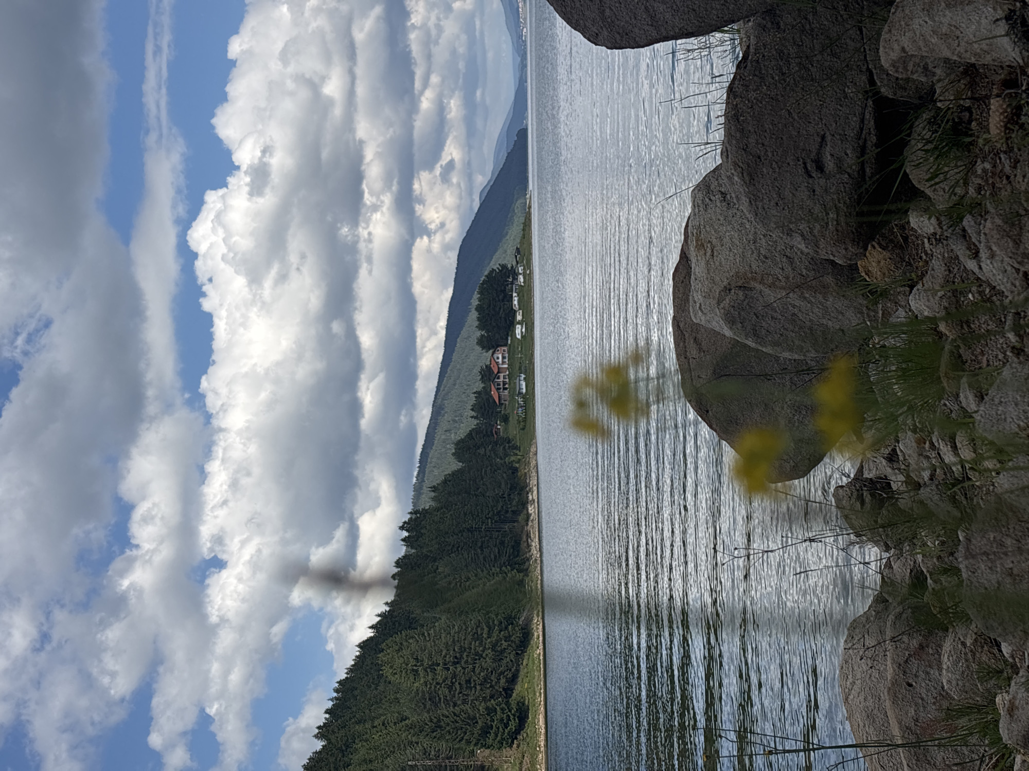 A body of water is in the foreground, with ripples on the surface reflecting the sky. Large rocks and sparse vegetation are visible along the shoreline. In the middle ground, a dense forest of evergreen trees covers a hillside that slopes down to the water. A few buildings, including what appears to be a house and some smaller structures, are nestled amongst the trees on the far bank. The background features rolling hills and a vast sky filled with dramatic clouds. A plume of dark smoke rises from the forest on the left side of the image.