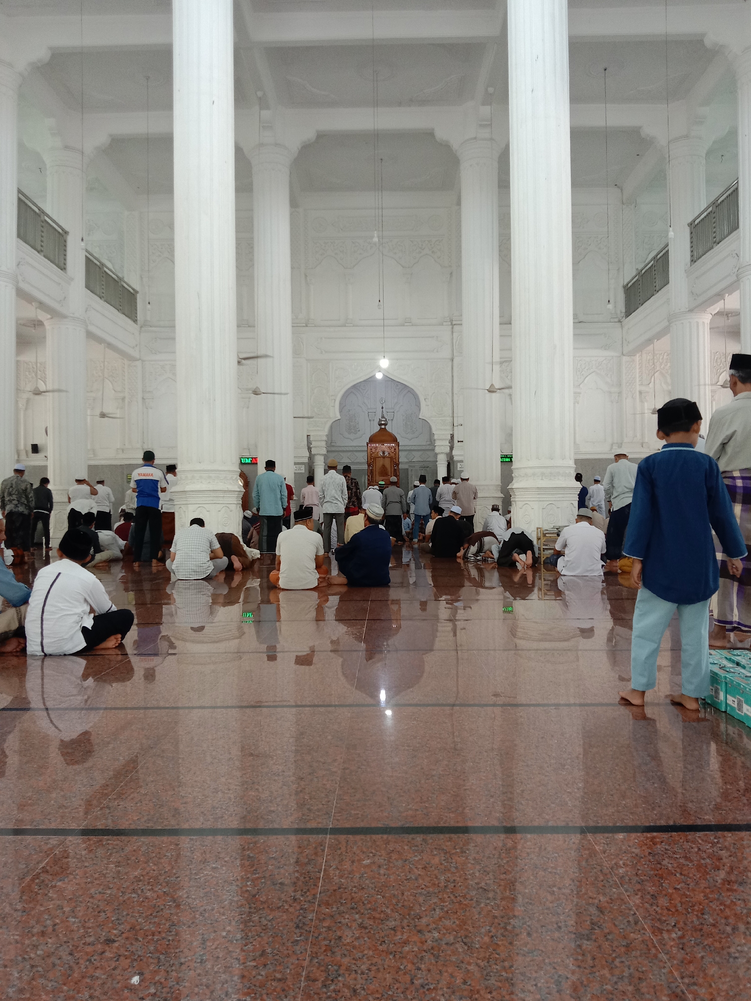 The image shows the interior of the Islamic Center Mosque in Lhokseumawe, Indonesia. A large group of Muslim men are engaged in prayer. Some are kneeling, some are prostrating, and others are sitting. The mosque features white architectural elements, including tall pillars and decorative walls. The floor is a glossy reddish-brown tile. A young boy in a blue shirt and light blue pants stands near the foreground, and other individuals are visible in the background, some wearing traditional Muslim attire.