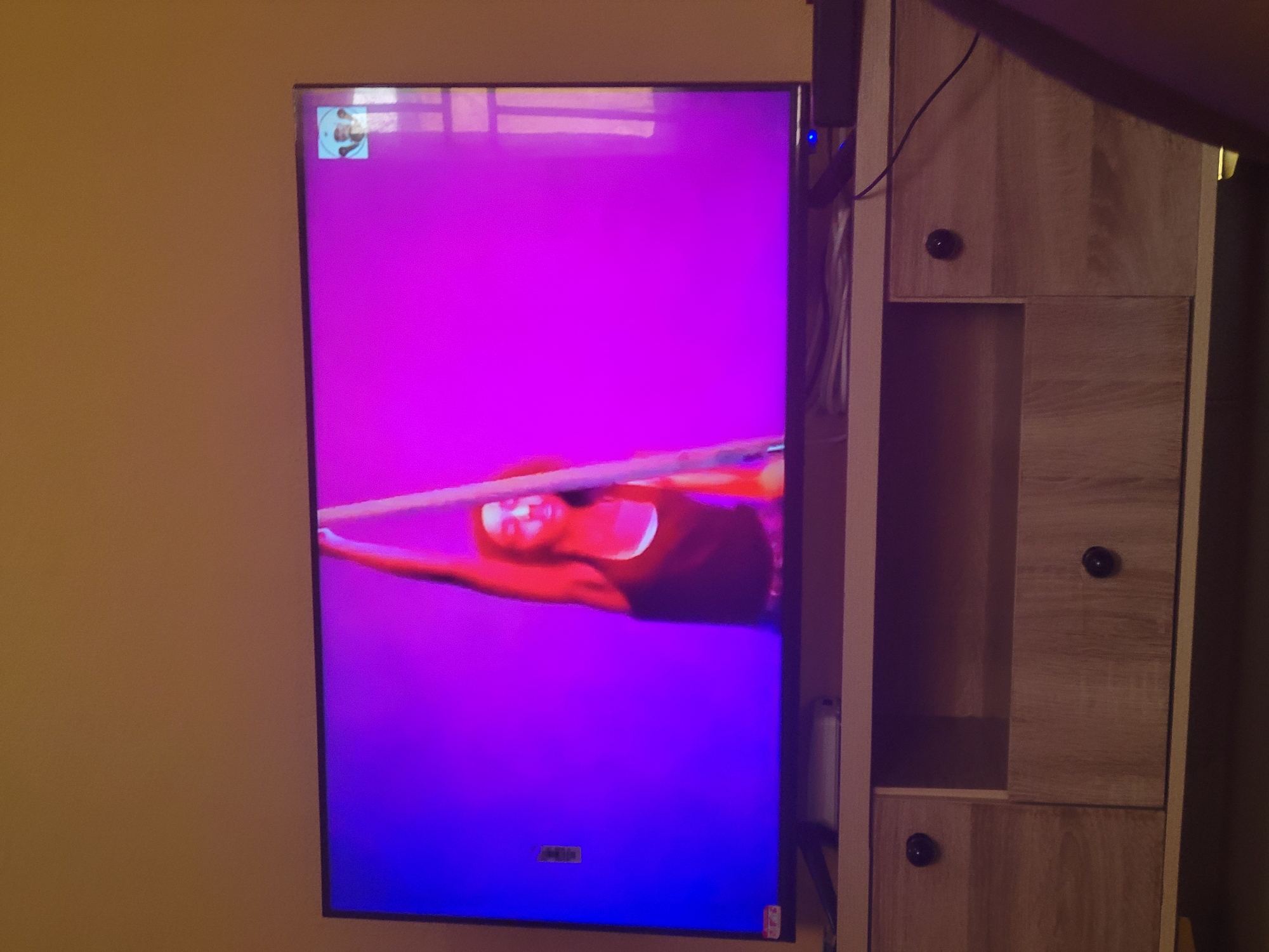 A television is mounted on a light brown wall, displaying a purple and blue image of a person performing aerial acrobatics on a suspended bar. To the right of the television is a light-colored wooden cabinet with black knobs. Wires are visible running from the television to the cabinet. The location is an indoor space in an unknown city.