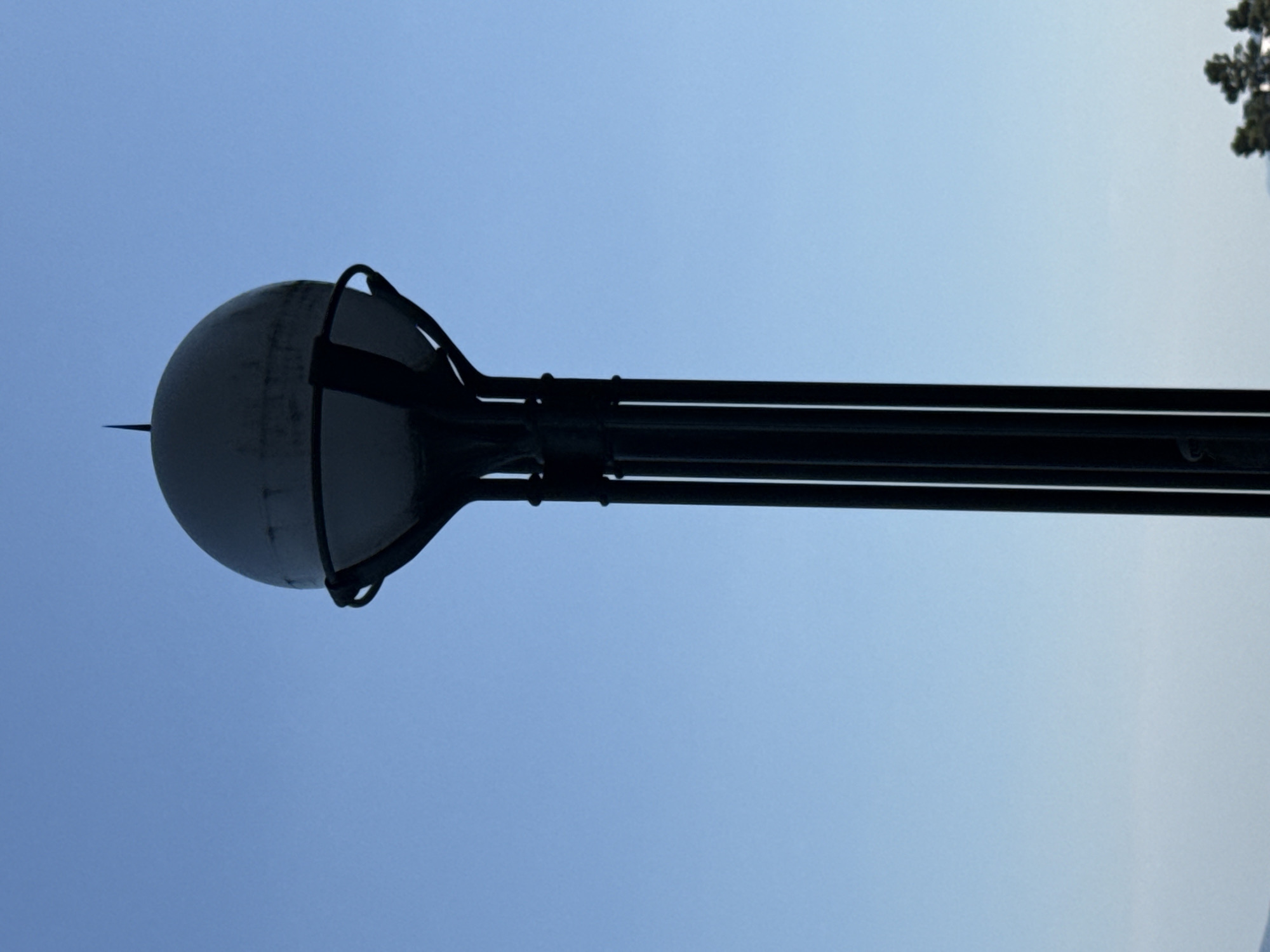 The image shows a tall, dark, pole-like structure topped with a spherical object. The structure is dark and appears to be made of metal, with several vertical supports. The spherical object is light-colored and has a pointed finial at its apex. The background is a clear, pale blue sky. The bottom of the image shows a sliver of blurry green foliage, suggesting the structure is outdoors. It is not possible to determine the specific location within the city of Opatija, Croatia from this image alone. There are no people or animals visible in the image.