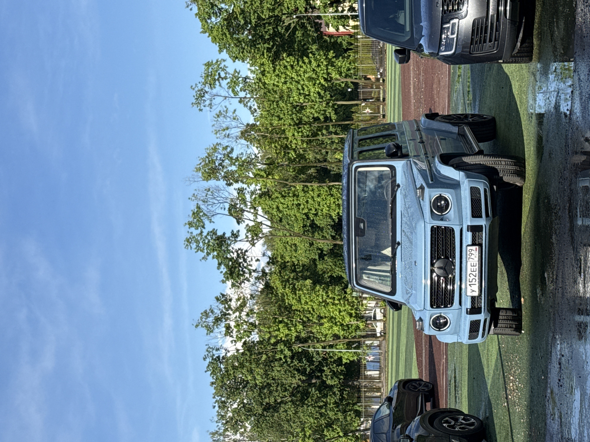 The image shows a light blue Mercedes-Benz G-Class parked on a green and brown surface, presumably artificial turf. The license plate reads "У 152 EE 799". Other cars are partially visible in the frame, including a dark-colored Range Rover at the top and the front of a black vehicle at the bottom. Trees fill the background, and a clear blue sky is visible above. Based on the license plate region code (799), the location is Moscow, Russia.