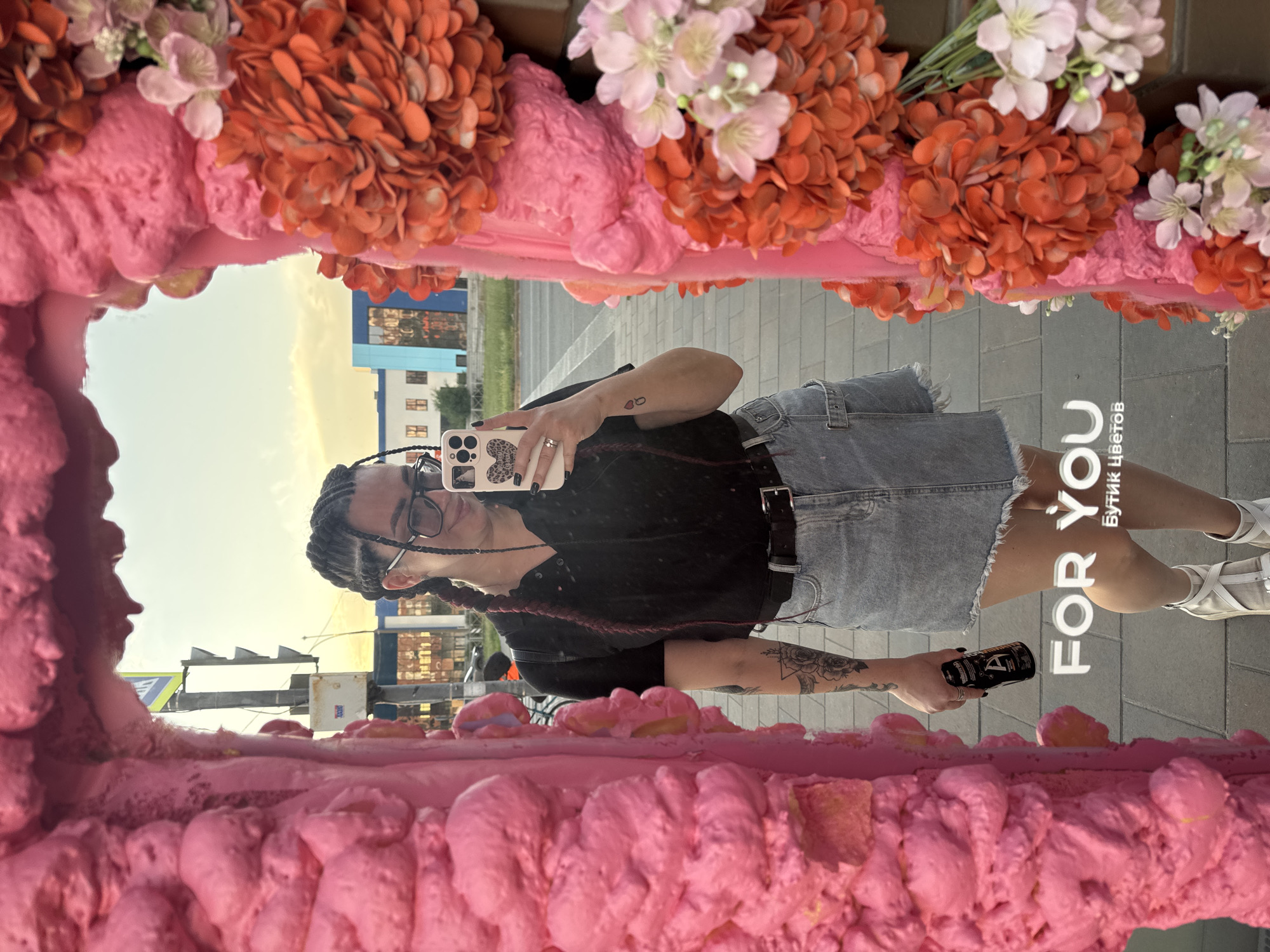 An outdoor, full-body mirror selfie is taken by a woman with dark braided hair and glasses, wearing a black shirt, denim skirt, and white sneakers. She is holding a black can in her right hand and a smartphone in her left. The mirror frame is decorated with pink foam and orange and pink faux flowers. In the background, buildings, a traffic light, and a street can be seen. Text overlay reads "FOR YOU" and "Бутик Цветов."