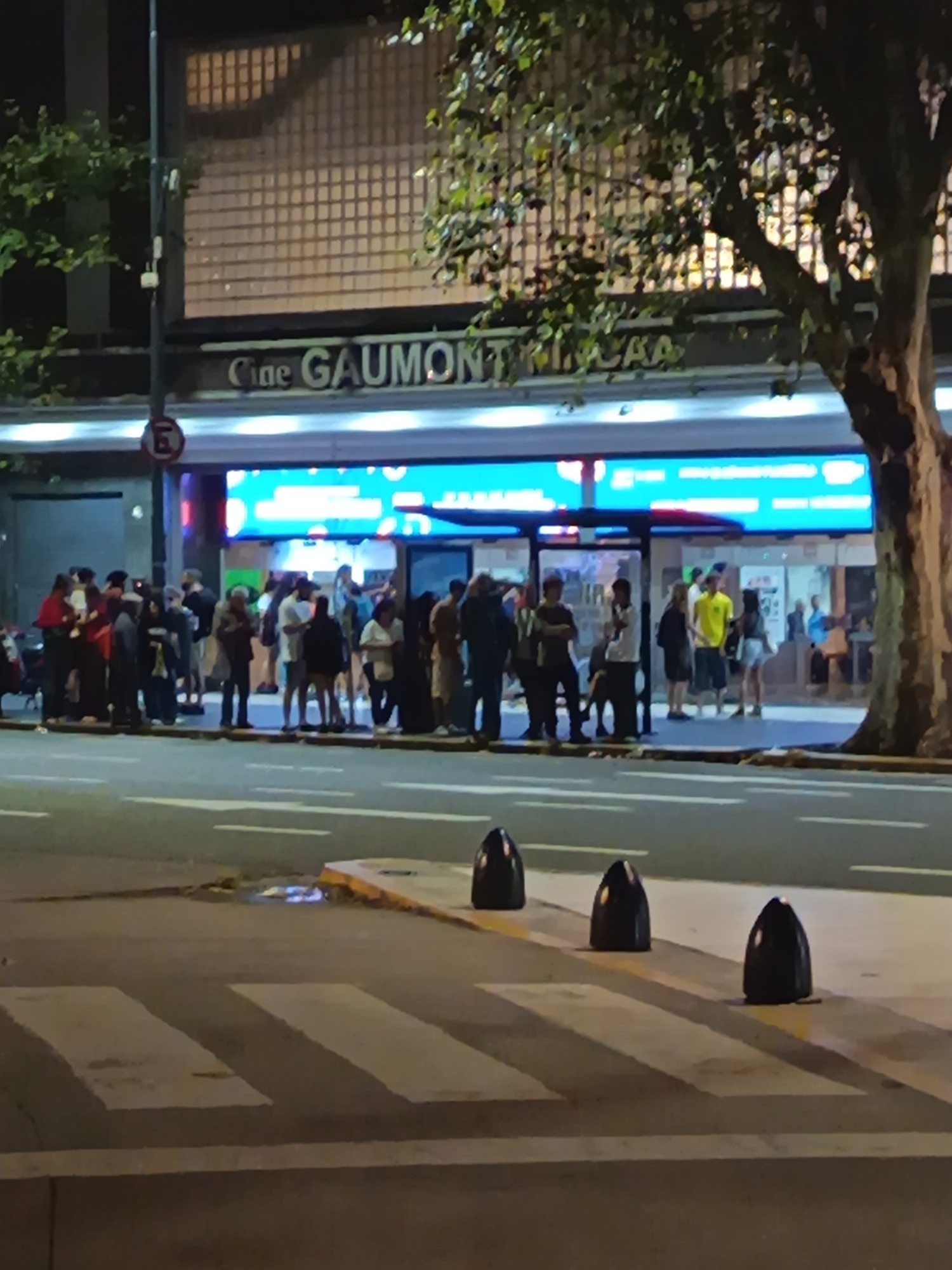 The image depicts the Cine Gaumont movie theater, located in Buenos Aires, Argentina. A group of approximately 30 people are standing in front of the theater, waiting in line. The theater's marquee is illuminated with a bright blue light. A large tree stands to the right of the theater entrance. A crosswalk is visible in the foreground, with black bollards separating the sidewalk from the street. A street sign with an "E" is visible on the left side of the picture.