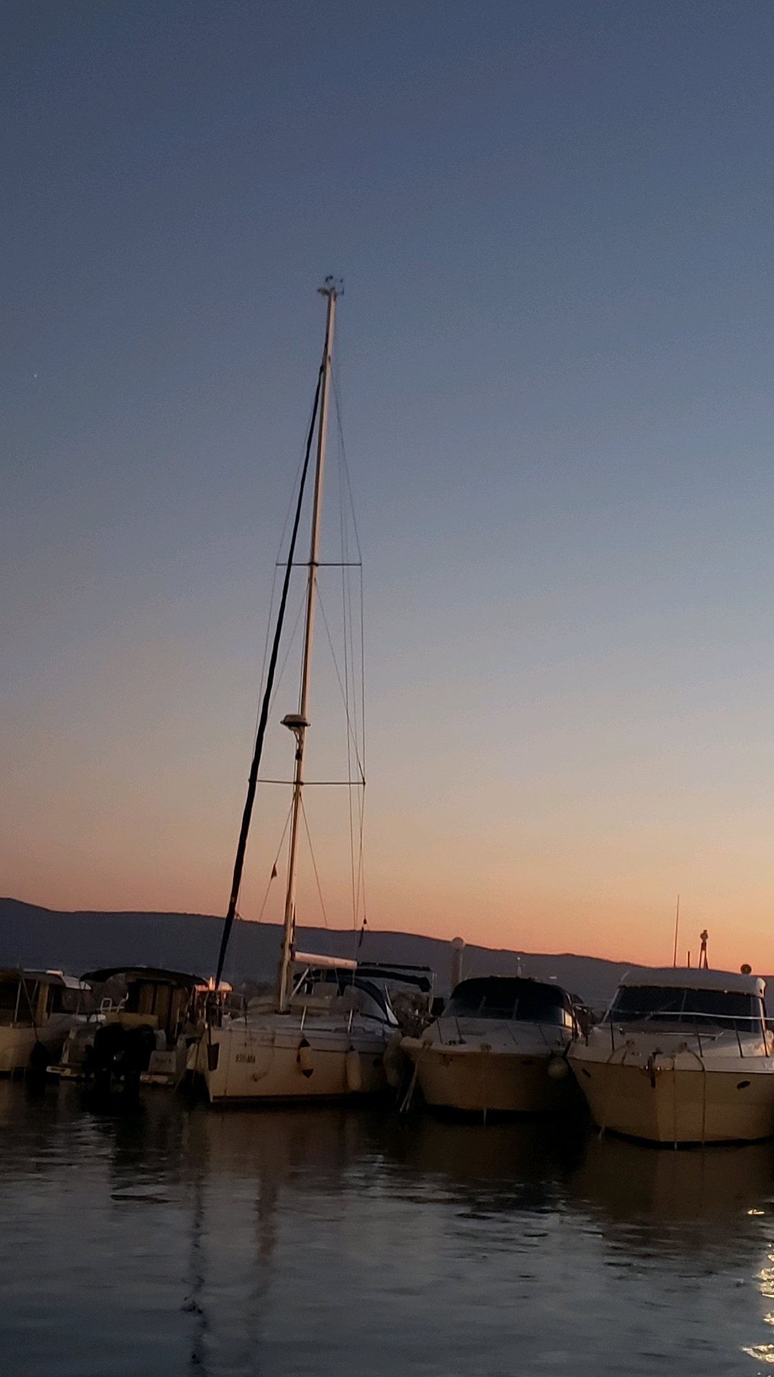 The image depicts a serene marina scene at dusk in Općina Tučepi, Croatia. Several boats are docked, including a prominent sailboat with a tall mast in the center, and several motorboats to its right. The water is calm, reflecting the soft hues of the twilight sky, which transitions from a clear blue at the top to shades of orange and pink near the horizon. In the background, a dark, silhouetted mountain range is visible. The overall atmosphere is peaceful and tranquil, characteristic of a marina at the end of the day.