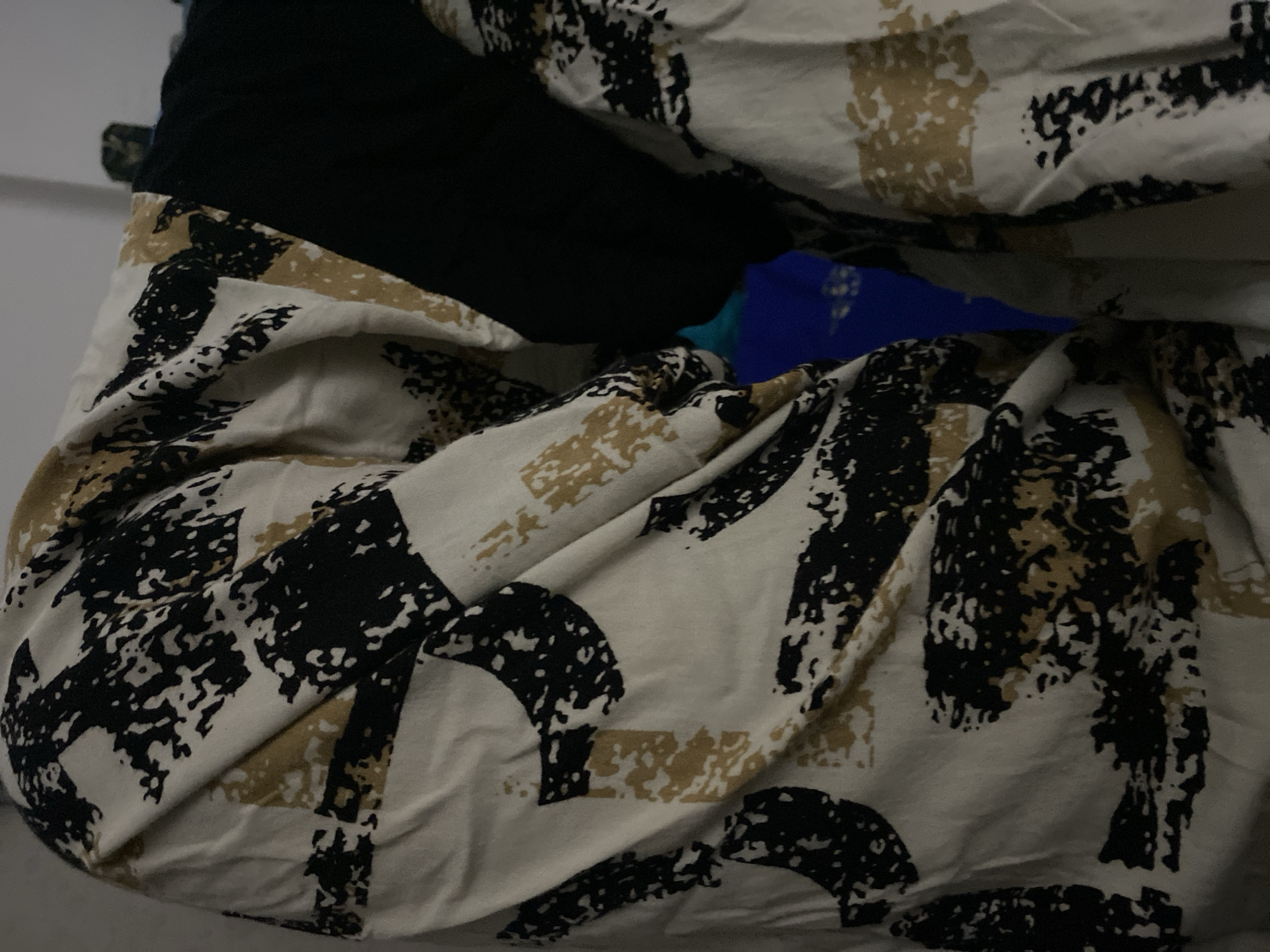 The image displays a close-up view of a patterned fabric, likely clothing, with an abstract design featuring black, tan, and off-white elements. The fabric is draped and creased, indicating it is flexible and possibly hanging or being worn. A section of plain black fabric is visible on the right side of the image, partially obscuring the patterned material and suggesting it might be a garment with contrasting panels or layers. In the background, a glimpse of a bright blue fabric or surface is visible. The lighting is dim, casting shadows that accentuate the texture and folds of the fabric. The scene appears to be indoors, possibly a domestic setting or a retail environment. There is no discernible information about the location in Ta', Indonesia.