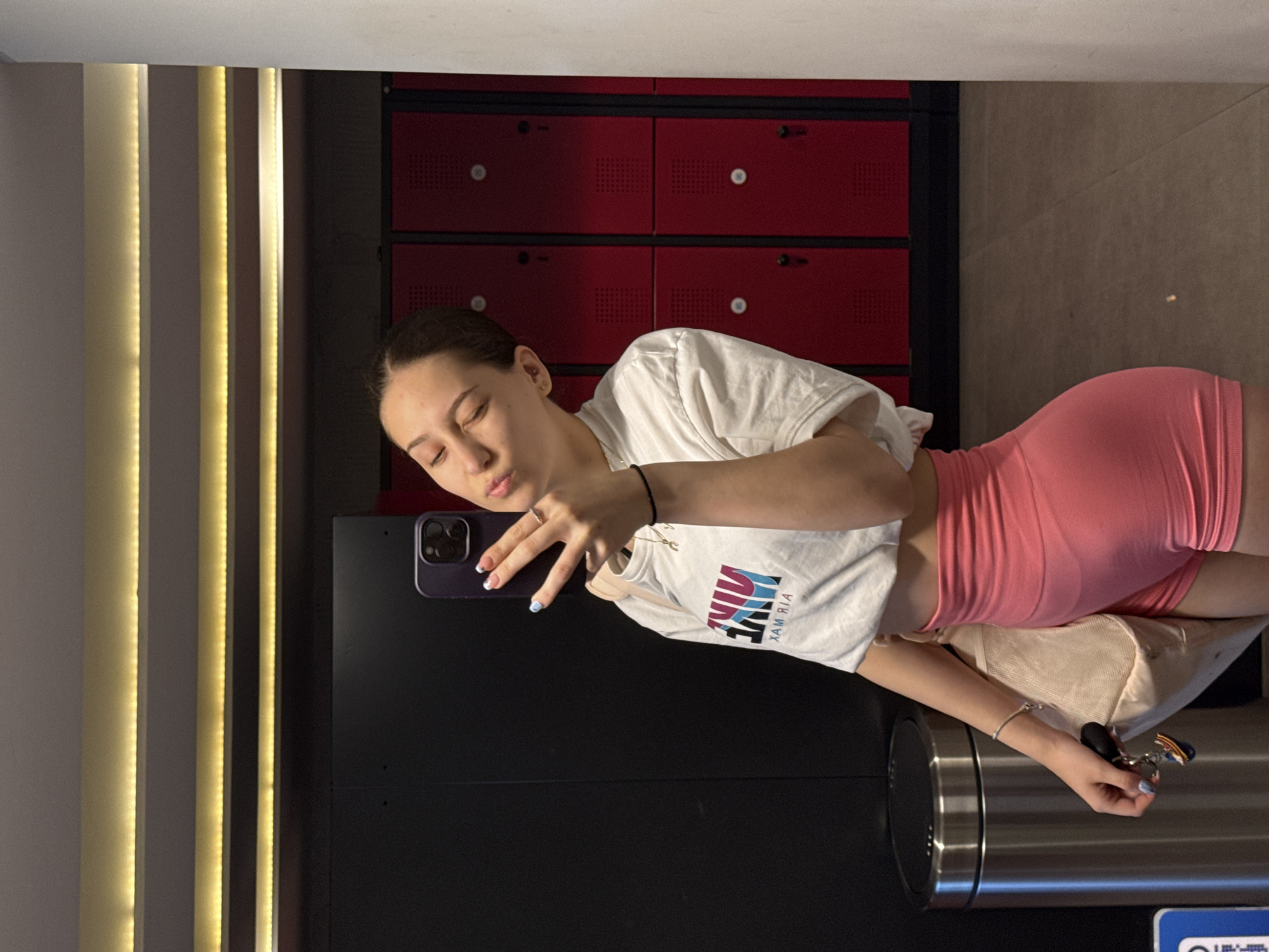 The image shows a young woman taking a selfie in what appears to be a gym locker room or similar facility. The background features a row of red lockers and another section with vertical gold-colored decorative lighting. She is wearing a white T-shirt with a "NIKE" logo, pink shorts, and accessories such as bracelets and a necklace. She is holding a purple phone with a multi-lens camera in her right hand, and her left hand is visible as well, exhibiting what looks like a manicured nail design. She is making a kissing face towards the camera. A metallic trash can and a beige bag are visible in the foreground. I am unable to confirm the specific location in Sofia, Bulgaria, based solely on this image.