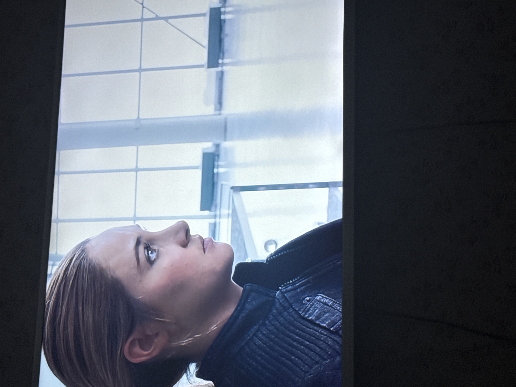 A medium shot captures a woman in a dark, quilted leather jacket, gazing upwards. The background reveals a modern building exterior featuring a grid of windows. Given the prompt's instruction to place the scene in Mytishchi, Russia, the architecture suggests a contemporary office or commercial building in the city. No specific actions or interactions are apparent.