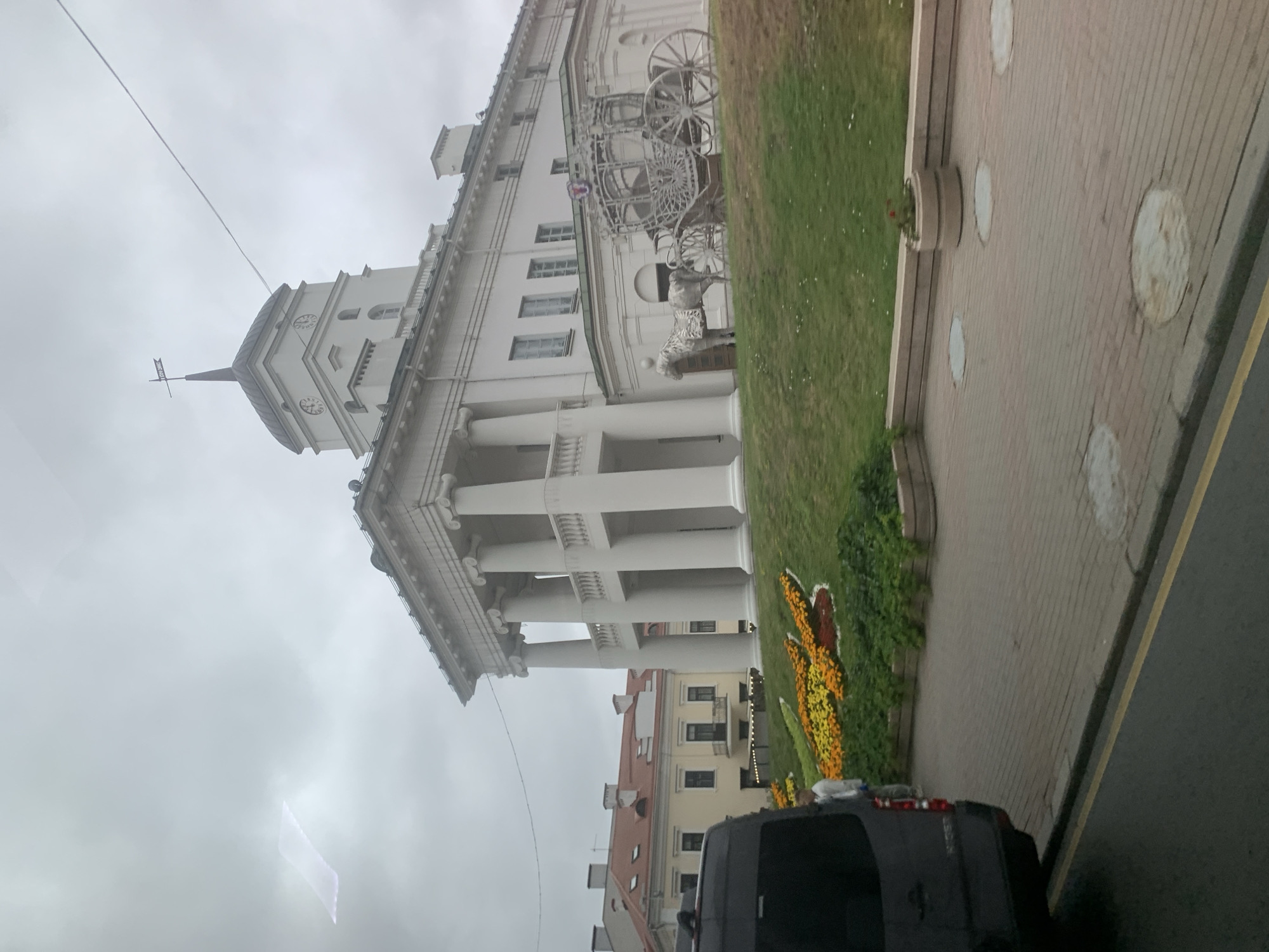 The image shows the Upper Town in Minsk, Belarus, specifically the City Hall building, which has a prominent clock tower topped with a weather vane. The building is white with classical architectural elements, including a colonnade with large columns. In front of the City Hall, there is a grassy slope with a colorful flowerbed shaped like a butterfly. A decorative horse sculpture is positioned near a carriage-like structure, which is likely part of an art installation or historical representation. The foreground shows a dark-colored van parked on the side of a road, and a paved walkway with circular manhole covers. The sky is overcast, suggesting a cloudy day.