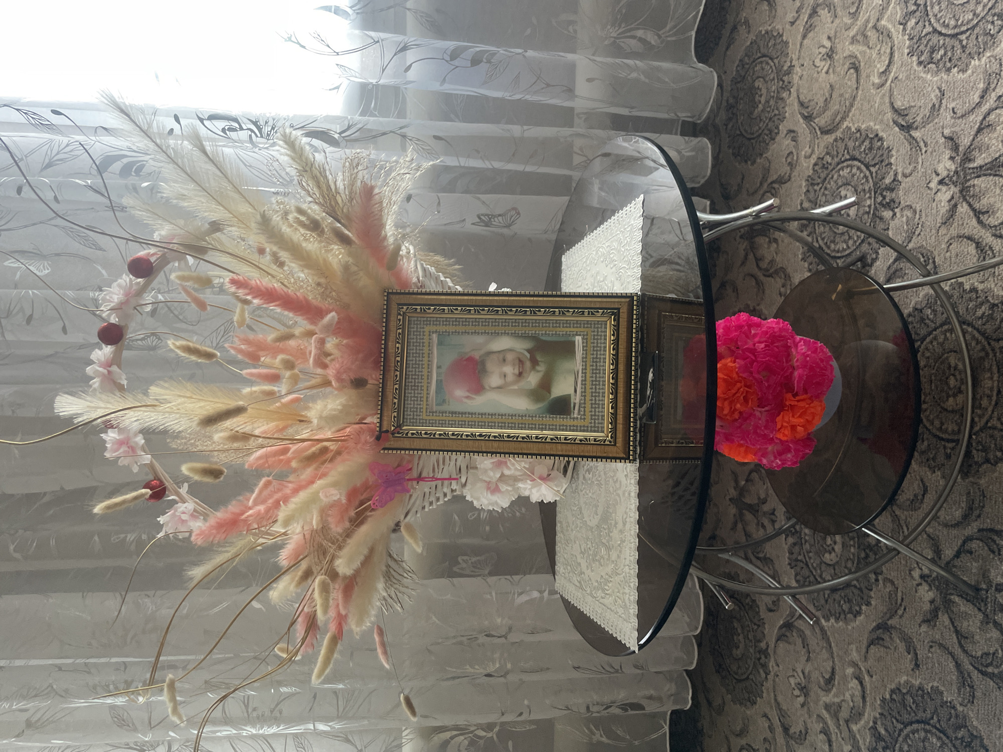 A framed photograph of a smiling child holding a red ball is placed on a glass-top coffee table. The photograph is in a decorative gold frame and is positioned in front of a large floral arrangement. The floral arrangement consists of dried pampas grass, bunny tail grass, and other fluffy, pastel-colored plumes in shades of pink and beige, arranged in a white wicker basket. Behind the arrangement, sheer curtains with a subtle leaf and butterfly pattern are visible. Below the coffee table, on a lower glass shelf, a small vase holds bright pink and orange flowers. The coffee table itself has a chrome-colored metal frame with curved legs. The scene is set against a carpet with a detailed, circular grey pattern. The lighting suggests a bright, possibly sunny day filtering through the curtains.