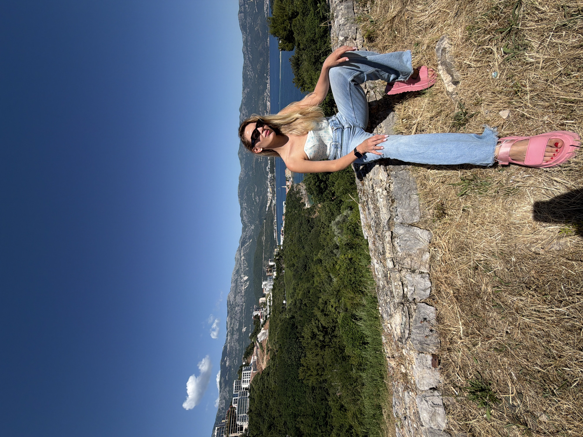 A young woman with long blonde hair and dark sunglasses is seated on a weathered stone wall, positioned at an elevated viewpoint overlooking the city of Budva, Montenegro. She is wearing a light blue strapless top with a subtle white pattern, light blue wide-leg jeans with frayed hems, and bright pink chunky sandals. A dark smartwatch is visible on her left wrist, and a ring adorns her right hand. Her legs are crossed at the ankles, with her left leg resting over her right, and her hands are placed on her thighs. She faces slightly to the right of the camera, looking towards it.
The background features a clear, deep blue sky with a few sparse white cumulus clouds. Below the horizon, rugged mountains, partially covered in green vegetation and dotted with modern white buildings (some appearing under construction), frame a deep blue bay. Within the bay, the distinctive red-roofed buildings of Budva Old Town are visible on a peninsula extending into the water, connected by a causeway. Dense green trees and bushes cover the hillsides between the elevated position of the woman and the bay. The immediate foreground consists of dry, golden-brown grass interspersed with small patches of green weeds, with shadows from the woman and the photographer visible on the ground.