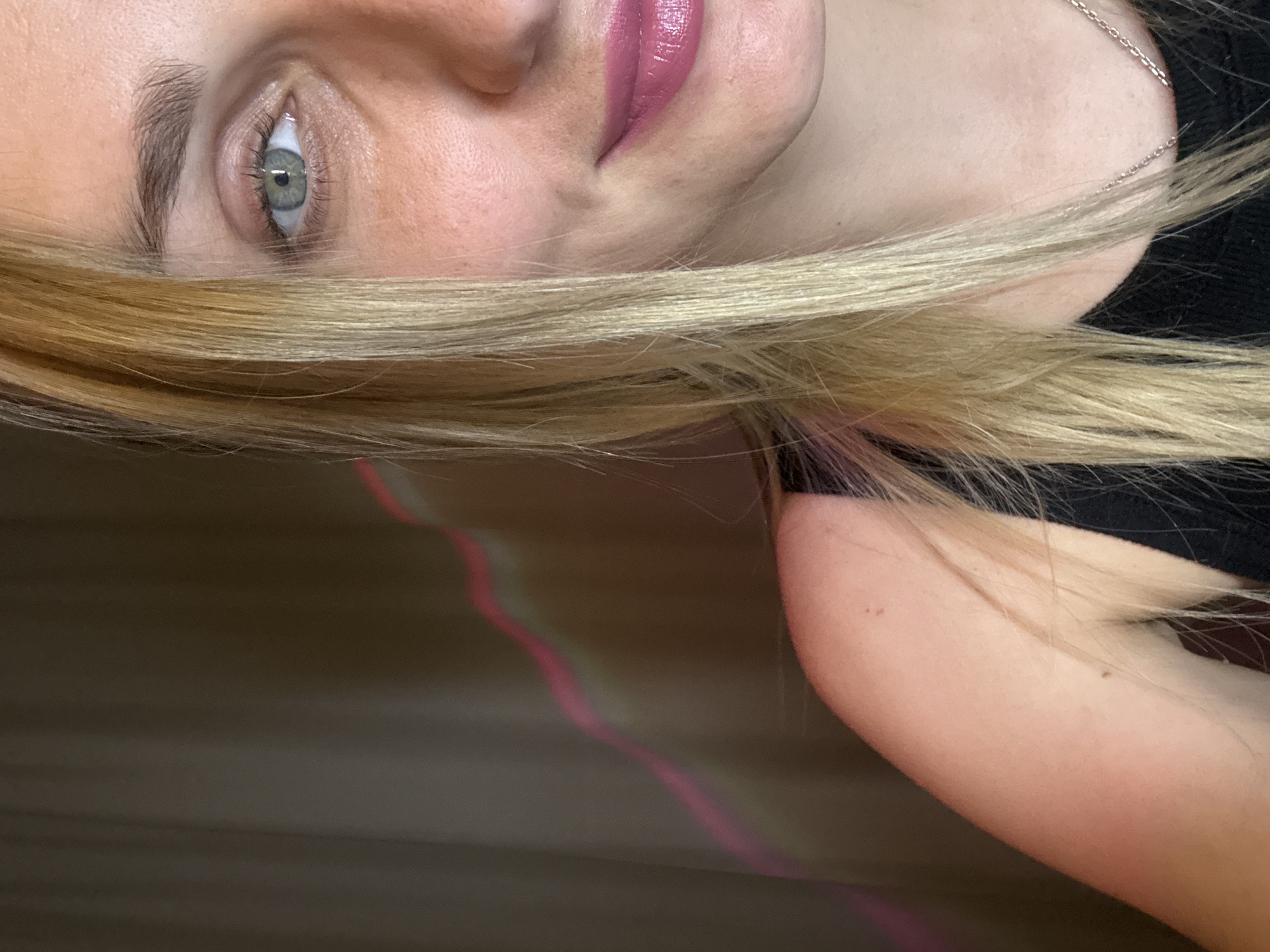 A close-up, high-angle shot shows the right side of a young, fair-skinned woman's face, from her eye to her shoulder. Her blonde hair is to the left of her face, with strands falling across her cheek. She has blue-green eyes and is wearing pink lipstick. Her skin appears smooth, with a faint dimple on her cheek. She is wearing a black top, and a silver necklace is visible. The background is a blurred, dark brown curtain with a pink wavy line of light running diagonally across it. The overall lighting is soft and diffused. The image is cropped and does not provide any information about the location.