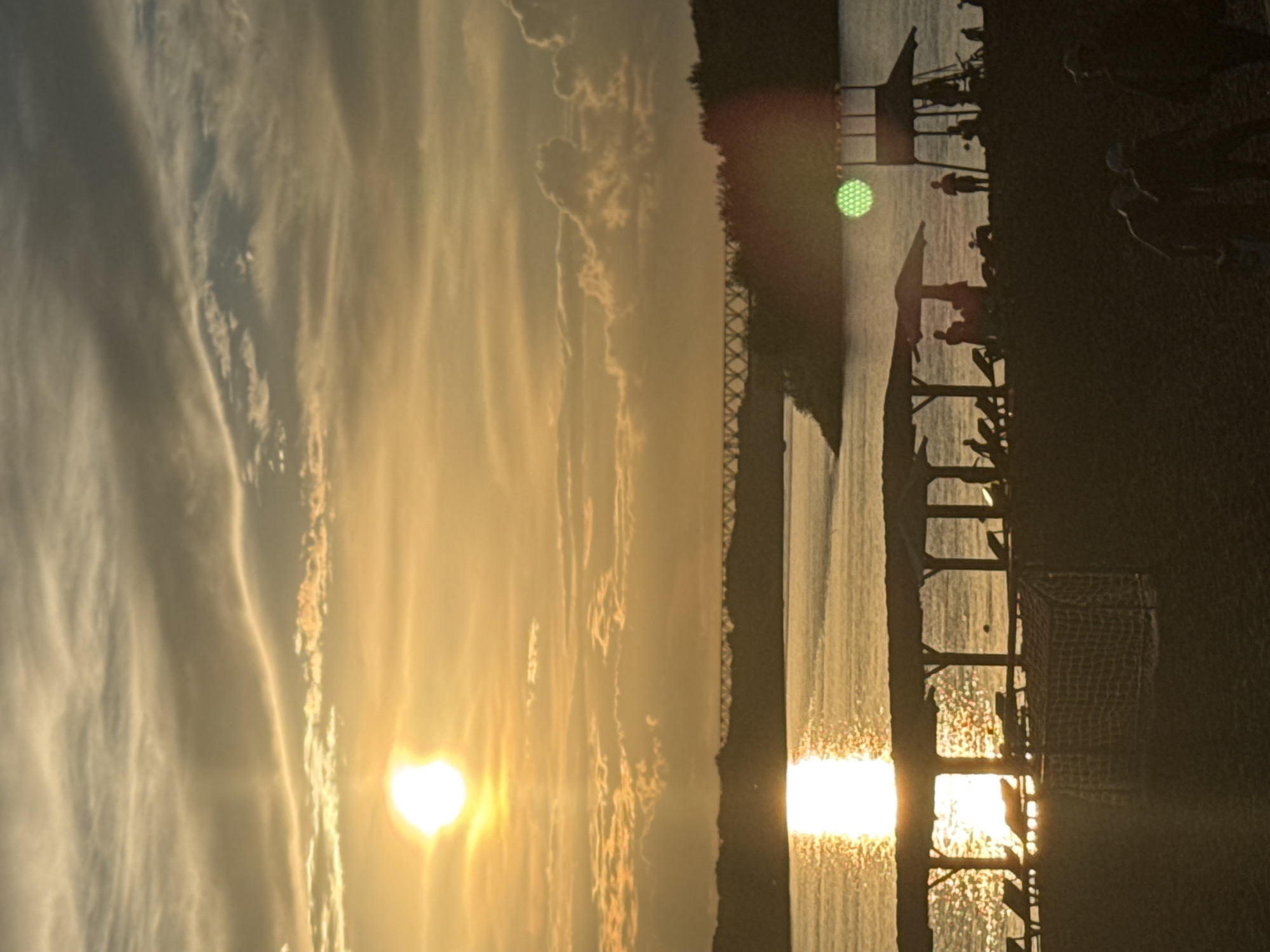 This is a photograph of a sunset over a body of water, likely a lake or river, given the location context of Pavlodar, Kazakhstan. The sky is filled with wispy, elongated clouds illuminated by the warm, golden light of the setting sun, which is prominently visible in the lower left portion of the frame. The water reflects the vibrant colors of the sky, creating a shimmering, textured surface.
In the foreground, silhouetted against the sunset, is a structure that appears to be a pier or a boardwalk. There are several figures of people on this structure, some appearing to be standing or walking, suggesting they are enjoying the view or perhaps engaged in some recreational activity. Two prominent umbrella-like structures are also visible on the pier, providing shade. Further to the right, a dark, textured area suggests land, possibly a beach or grassy area, and a soccer goal is partially visible. A faint green, circular light is also present on the pier structure.
The overall mood is serene and peaceful, with the warm hues of the sunset evoking a sense of tranquility and the end of a day. The presence of people suggests a public space, possibly a park or recreational area. The time of day is clearly sunset, characterized by the low angle of the sun and the dramatic lighting. The weather appears clear, with the clouds adding a dynamic element to the sky. There is no visible text in the image.