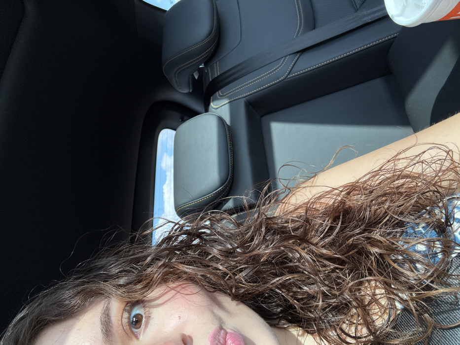 A selfie in a car shows a young woman with wet, curly brown hair and brown eyes. Her lips are slightly pursed. The car's interior is visible, featuring black leather seats with light stitching and a safety belt. A window provides a glimpse of the sky with light clouds. A drink container is visible in the top right corner.