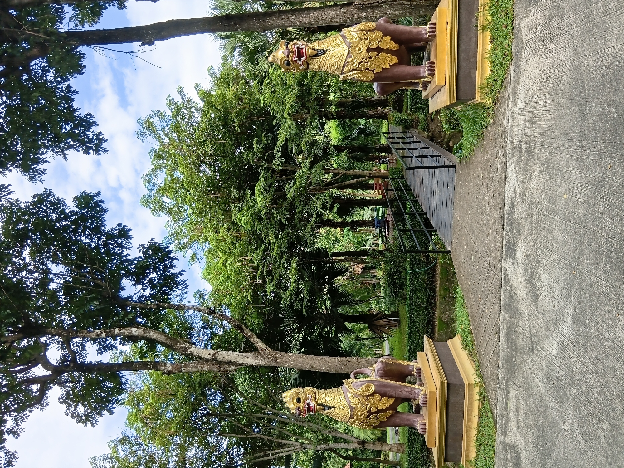 This image depicts a park in Ban Pa Phai Si Khong, Thailand, featuring two large, ornate statues of mythical lion-like creatures. These statues, made of a dark material with elaborate gold ornamentation, are positioned on pedestals on either side of a pathway. The pathway leads to a wooden bridge that spans a small body of water, surrounded by lush green foliage and mature trees. The sky above is partly cloudy. In the background, a person can be seen walking across the bridge. The overall environment suggests a tranquil, culturally significant outdoor space.