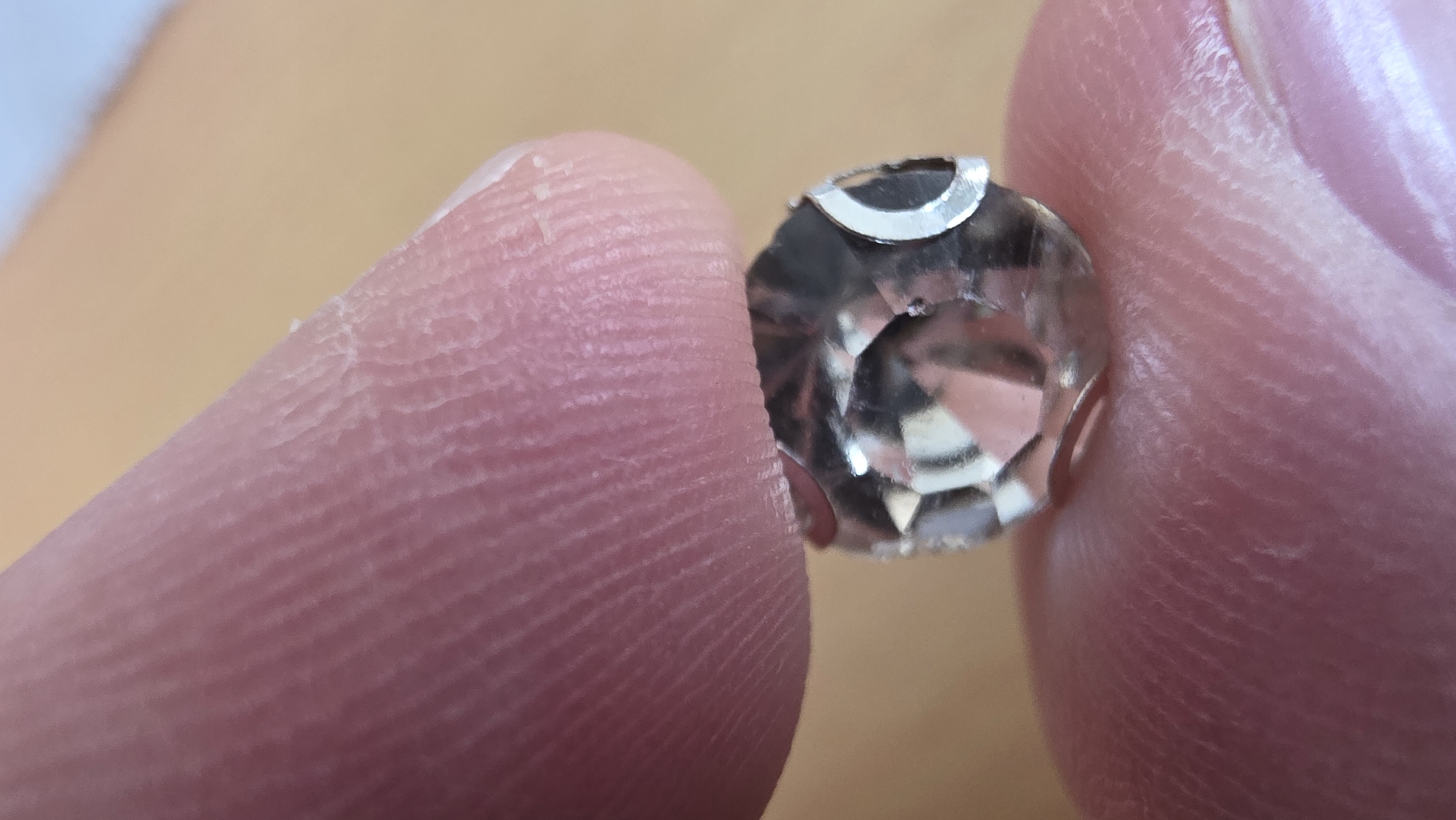 A close-up, macro photograph captures two human fingers gently holding a small, clear, faceted object. The fingers, likely belonging to one individual, are positioned to present the item centrally. The skin texture of the fingers is highly detailed, revealing distinct fingerprints, fine lines, and subtle creases; some areas appear slightly dry or textured.
The object itself is transparent and features numerous reflective facets, indicative of a cut gemstone, crystal, or decorative bead. A notable detail is a metallic, C-shaped clip or setting visible on the top edge of the object. The various facets brilliantly reflect ambient light, highlighting the object's polished surface.
The background is intentionally out of focus, rendered in soft, warm tones of light brown and beige, suggesting an indoor setting with a neutral surface or diffused lighting. There are no other discernible objects or specific environmental cues in the blurred background. The primary activity depicted is the careful holding and display of the small, reflective item. No text is visible within the image, and due to the absence of faces or broader context, no emotions, specific time of day, or weather conditions can be inferred.