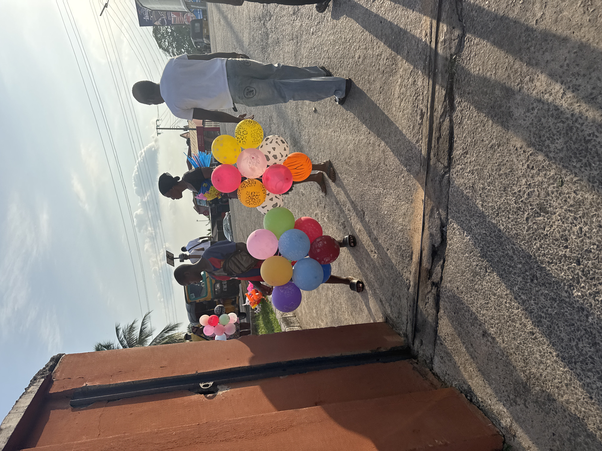 A street scene in Onitsha, Nigeria, shows a barefoot child carrying a large bundle of balloons of various colors and patterns. The child walks on a concrete sidewalk, partially obscured by the shadow of a nearby structure. An adult in a white shirt and gray pants is walking ahead of the child. A building with an orange-brown exterior is in the foreground, with a glimpse of additional balloons displayed nearby. Other pedestrians and vehicles are visible in the background, with overhead power lines and a partly cloudy sky.