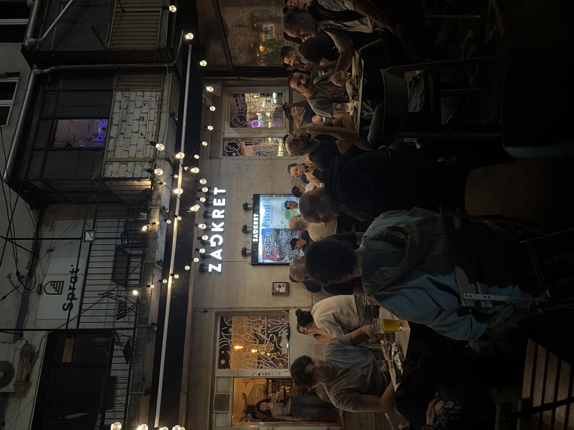 The image captures an outdoor seating area of a establishment in Belgrade, Serbia, at night. The sign above reads "ZADKRET". String lights illuminate the scene, casting a warm glow on patrons seated at various tables. The architecture suggests an urban setting, possibly an alleyway or courtyard.
In the foreground, several individuals are seen dining and socializing. A man in a grey t-shirt is seated at a table with a glass of beer and a foil-wrapped item. To his right, a person in a light blue hooded jacket is facing away from the camera, seated on a chair. Other patrons are visible in the middle ground and background, engaged in conversations and enjoying their evening.
The establishment appears to be a bar or restaurant, with windows revealing interior lighting and decorations. A woman carrying a tray is seen entering through a doorway on the left. The overall atmosphere is relaxed and convivial, typical of an evening gathering at an outdoor urban venue.