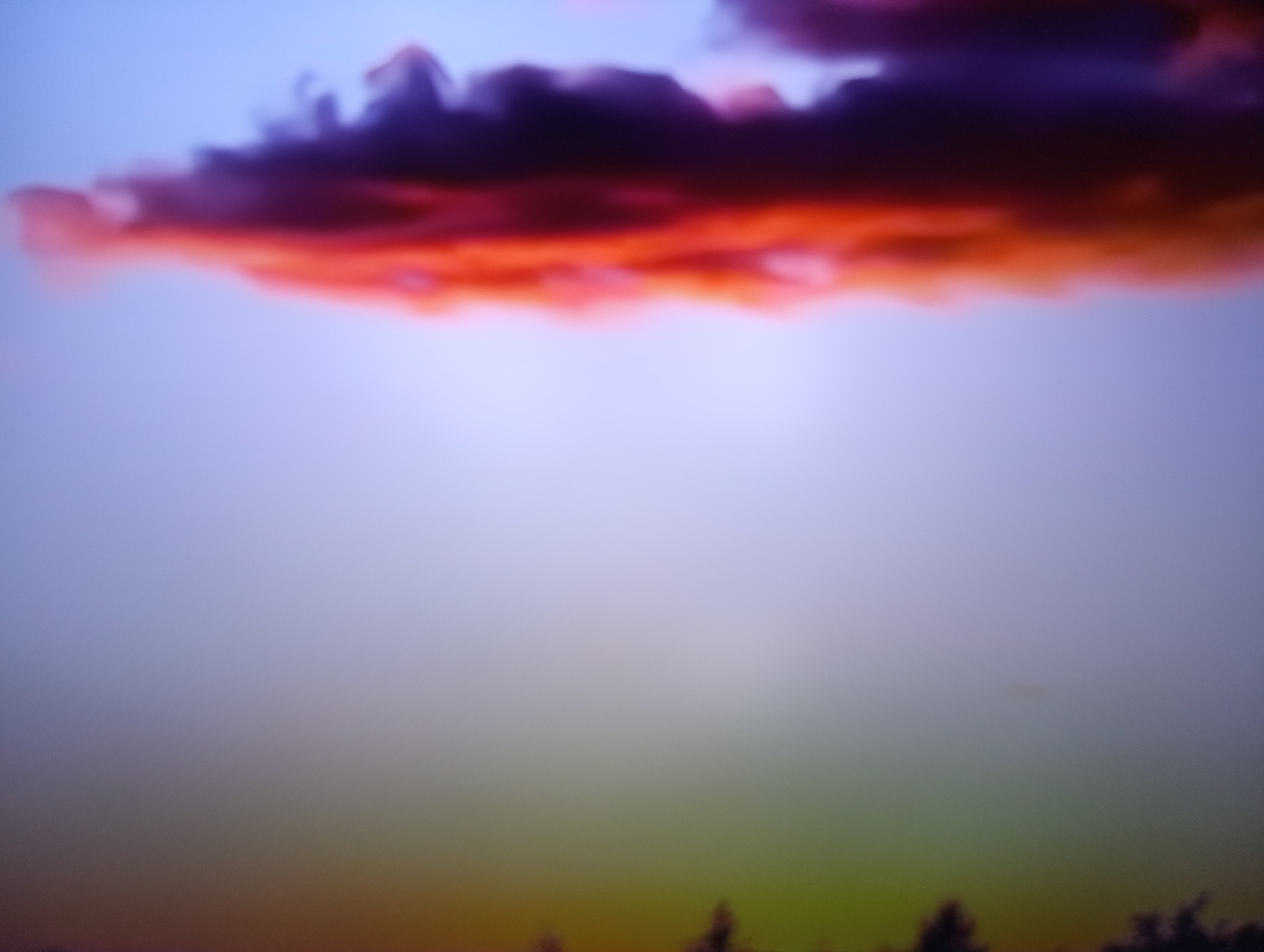 This is a blurred, abstract photograph of a sunset or sunrise. The primary subject is a dramatic cloud formation in the upper portion of the frame, colored in intense shades of orange, red, and deep purple. The cloud appears to be illuminated from behind by the setting or rising sun.
Below the cloud, the sky transitions through soft purples and blues, fading into a hazy, greenish-yellow hue at the bottom of the image. This lower section suggests the presence of a landscape, possibly silhouetted trees or foliage, though they are indistinct due to the blur and low light. The overall mood is ethereal and visually striking, emphasizing color and light over sharp detail.
The time of day is clearly indicated as either dawn or dusk by the quality of light and the vibrant sunset/sunrise colors. There are no visible people, text, or specific objects that provide precise location cues beyond the general suggestion of a natural landscape. The emotional tone evoked is one of awe and tranquility, stemming from the beautiful, if abstract, depiction of the sky's natural spectacle. The location context of Dnipro, Ukraine, places this natural phenomenon within that geographical setting.
