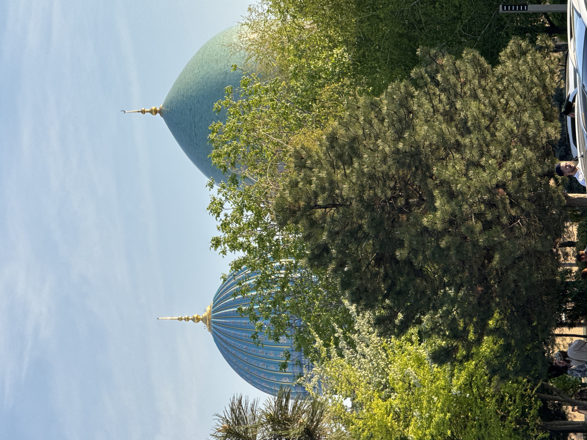 The image captures a view of two domes of a building in Tashkent, Uzbekistan. The first dome has a green color, and the second has a blue color. The domes are embellished with golden spires. The foreground features green trees, partially obscuring the lower portions of the domes. A tour bus with people is visible on the right edge of the frame. The background consists of a clear, pale blue sky.