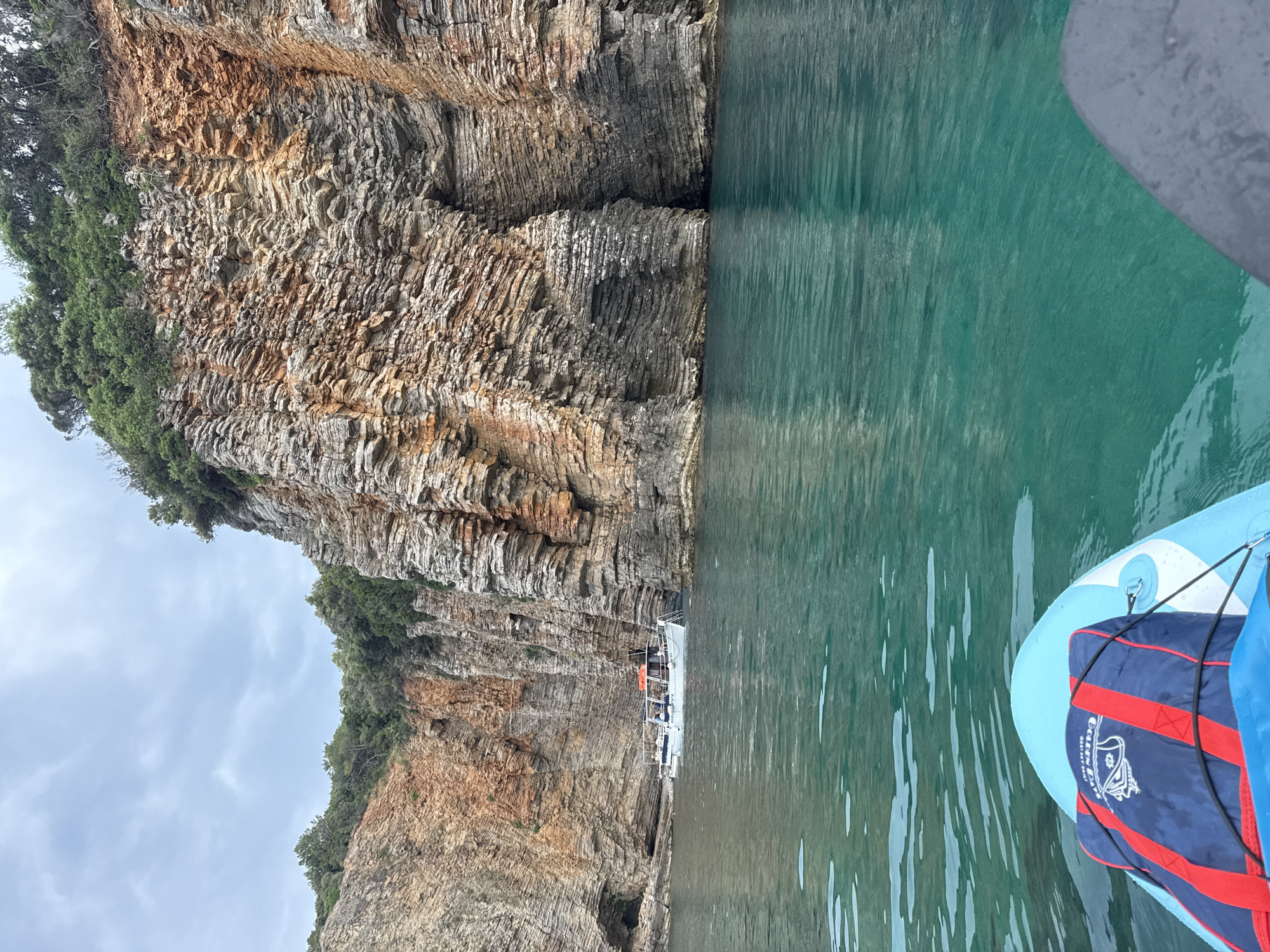 The image shows a cliff face with a body of water in the foreground. A stand-up paddleboard with a life vest is visible on the right side of the frame. The cliff is rocky and covered with some vegetation at the top. The water appears to be a calm, turquoise color. In the distance, a boat is moored near the cliff face. The sky is overcast, suggesting it could be a cloudy day. The scene evokes a sense of outdoor adventure and exploration in a coastal environment. The life vest on the paddleboard has text on it: "GOLDEN DA".