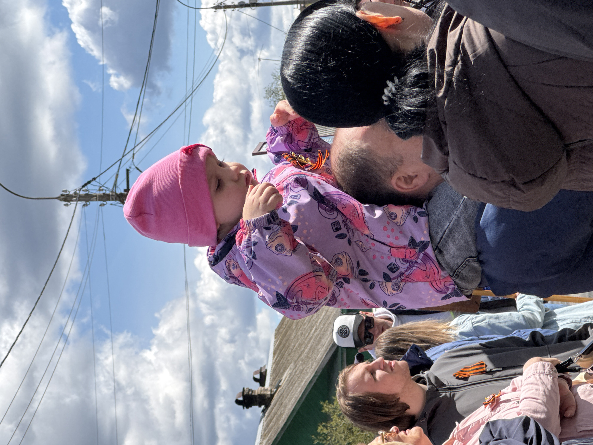 The image captures a scene outdoors on a bright day, likely in Yamyshevo, Russia, given the St. George ribbon visible on some of the individuals. The central subject is a small child wearing a pink beanie and a patterned purple jacket, held up by an adult. The adult holding the child appears to be bending over slightly to allow the child to interact with another person, possibly a family member, who is standing to the side. A woman with black hair is positioned close to the adult holding the child. Several other people are visible in the background, including individuals wearing jackets and casual clothing. Some wear sunglasses. Overhead, there are power lines visible against a partly cloudy blue sky. In the midground, there is a building with a green roof.
