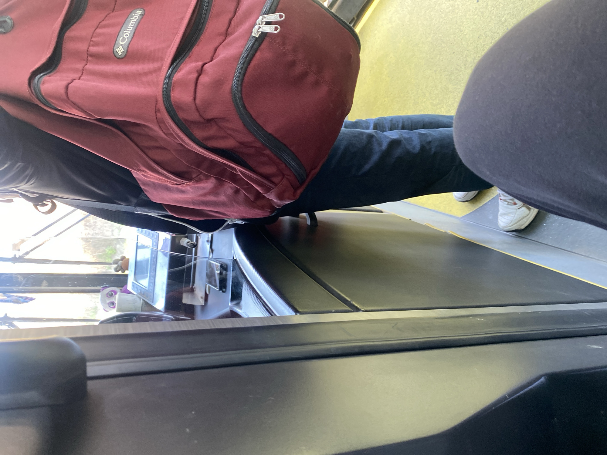 The image shows the interior of what appears to be a bus or other public transportation vehicle. In the foreground is a section of the vehicle's structure, likely part of the dashboard or a seating partition, with a black, glossy surface. Further back, there is a passenger with a dark red Columbia backpack with black zippers. The passenger is wearing dark jeans and white athletic shoes. Another person in black pants sits on the right of the frame. Outside the window, in the background, is a glimpse of the exterior world, though details are obscured. There is no information regarding the location being in Varna, Bulgaria in the given image. The objects on the shelf near the window appear to be a small television screen with a small plush toy and other small objects.