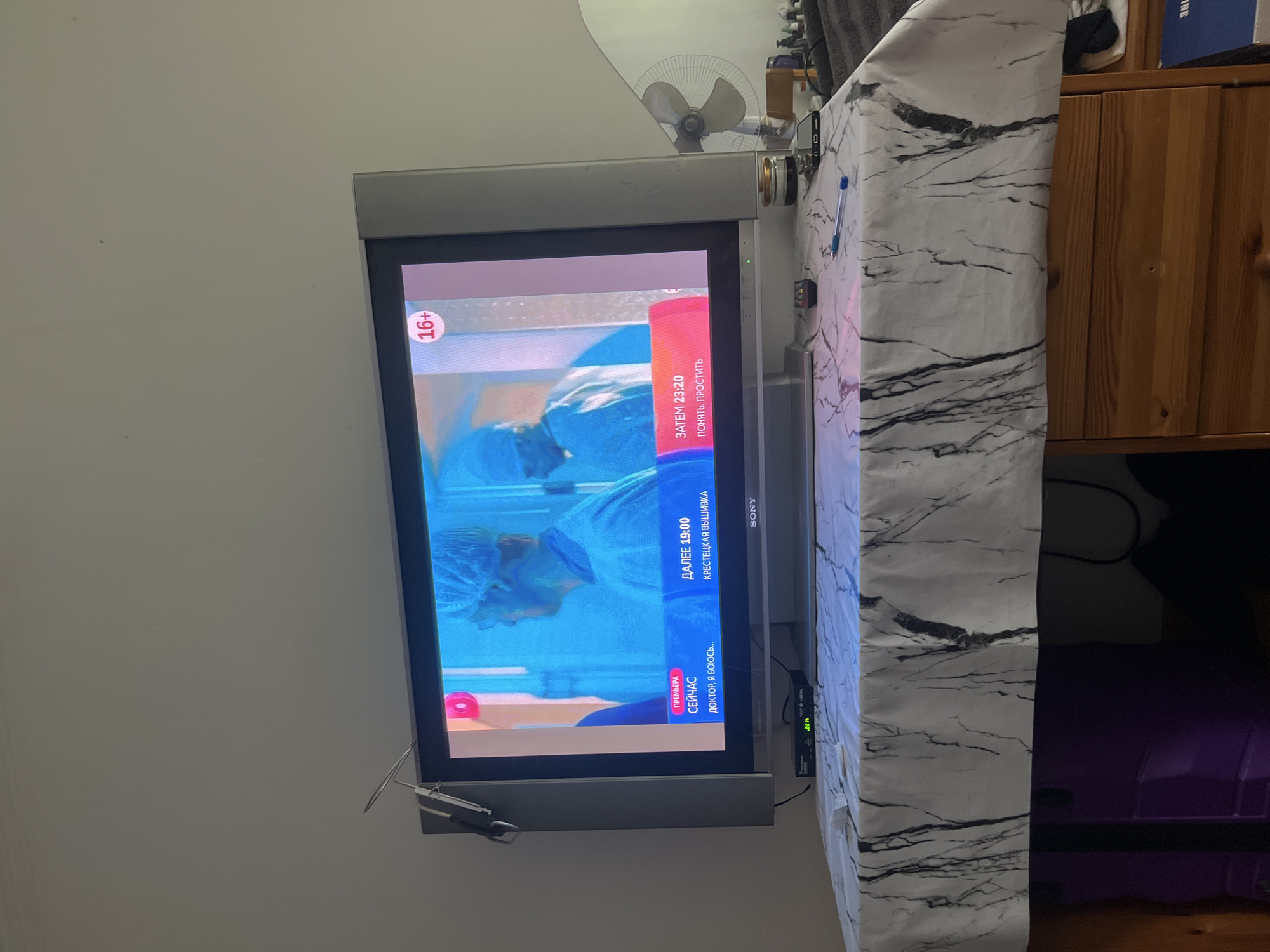 The image shows a television screen displaying what appears to be a program with Russian text. The television is a flat-screen model, likely a Sony, placed on top of a dresser covered with a marble-patterned tablecloth. The dresser is made of light-colored wood and has drawers. On the dresser, a black set-top box is visible below the TV, along with various small objects including a remote control, a smartphone, a pen, and what seems to be a small container. To the right of the television, a white fan is positioned. The background is a plain, light-colored wall, and a portion of a mirror is visible to the right of the fan. The overall environment suggests a domestic setting. There are no individuals clearly visible in the scene. The program on the screen displays text such as "ПРЕМЬЕРА", "СЕЙЧАС", "ДАЛЕЕ 19:00", and "ЗАТЕМ 23:20", indicating it is a TV schedule or broadcast information, with the age rating "16+" also displayed. The text "ДОКТОР, Я БОЮСЬ..." and "КРЕСТЦОВАЯ ВЫШИВКА" are also visible, suggesting program titles or descriptions. The scene is indoors, likely in a living room or bedroom. The location of Nalchik, Russia, cannot be determined from the visual information present in the image.