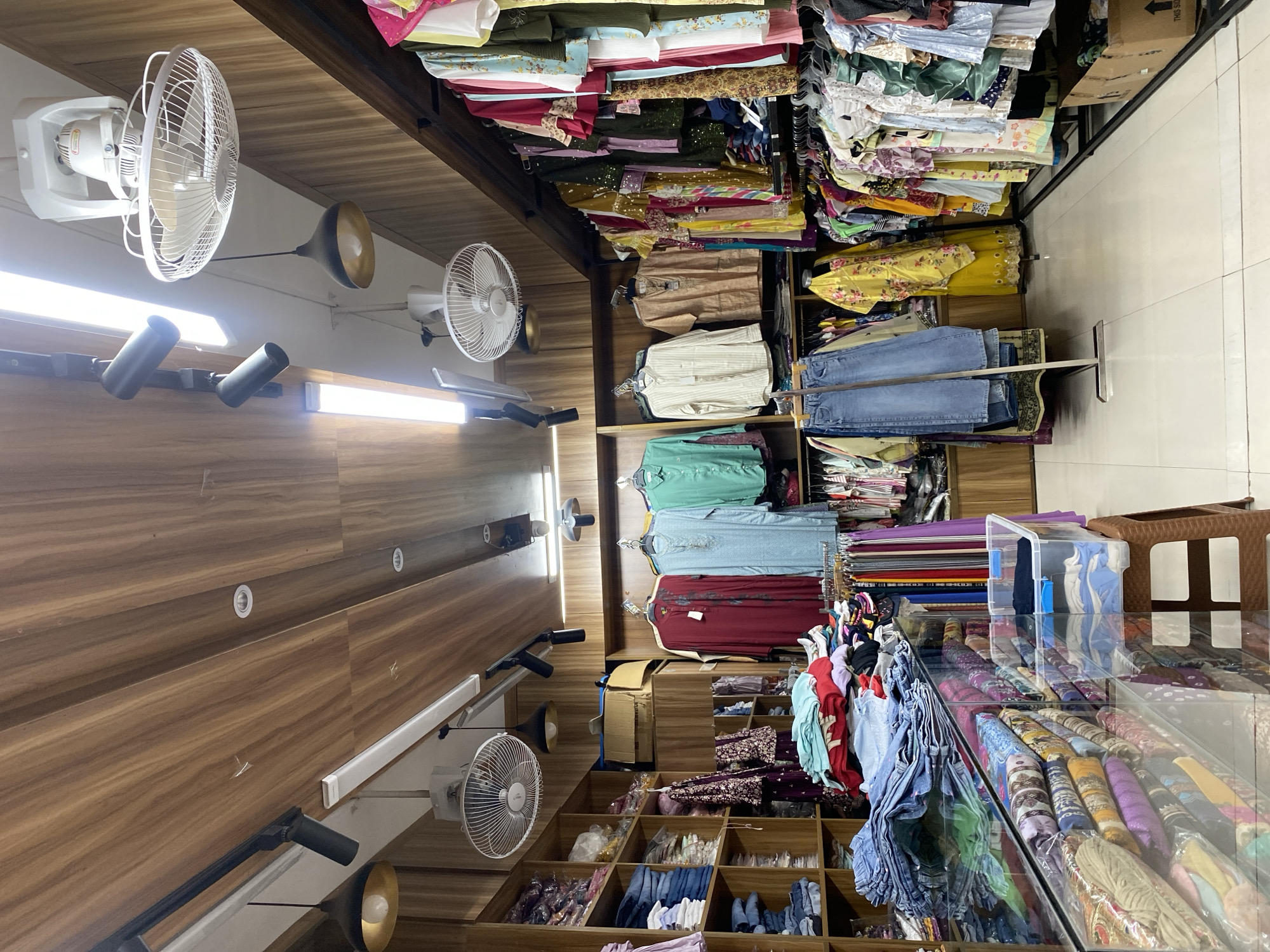 The image depicts the interior of a clothing store in Dhaka, Bangladesh. The store features wooden paneled walls and ceiling, with various clothing items displayed on shelves and racks. Several ceiling fans and track lighting fixtures are visible. On the left, shelves are stocked with folded jeans and other garments. In the center, a rack holds several kurtis (traditional tunics) in colors like maroon, light blue, and green. To the right, a display rack showcases a variety of colorful women's clothing, including dresses and kurtis. In the foreground, a glass display case contains folded textiles. A plastic storage bin with folded clothes sits on a small stool. The store appears well-stocked with a diverse range of apparel.