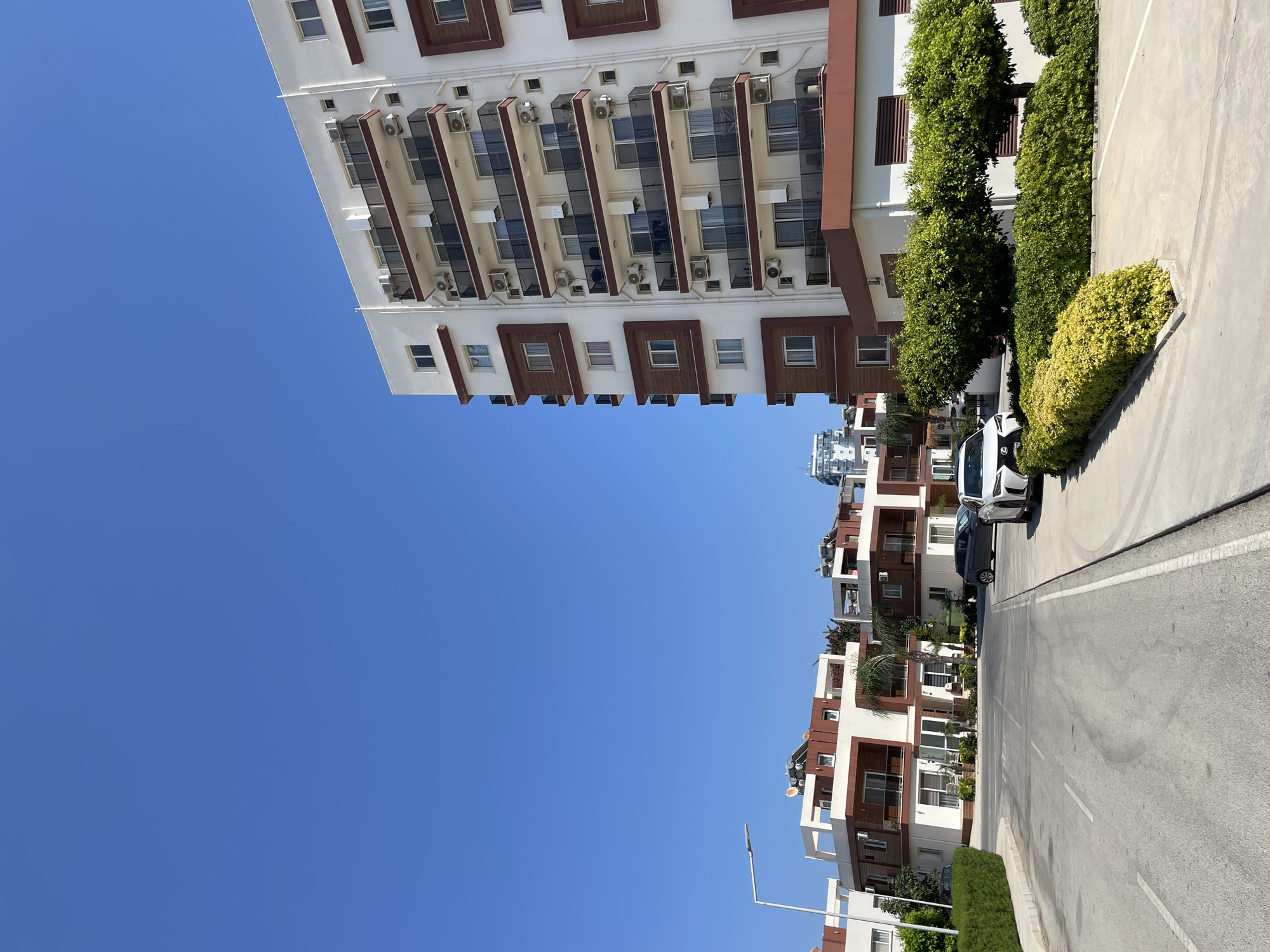 The image shows a residential area in Trikomo, Northern Cyprus/Cyprus, under a clear blue sky. In the foreground, a paved road curves to the left, bordered by a sidewalk and neatly trimmed bushes. To the right of the road, a modern white apartment building with brown accents stands tall, featuring multiple balconies with dark railings and numerous air conditioning units. A row of smaller, multi-story residential buildings with similar architectural style and brown trim is visible further down the road to the left. Several cars are parked along the road and on the properties. Lush green trees and manicured shrubbery are interspersed throughout the scene, adding to the well-maintained appearance of the neighborhood. In the background, a glimpse of a taller, modern building can be seen beyond the residential structures. The overall impression is that of a sunny, contemporary residential development.