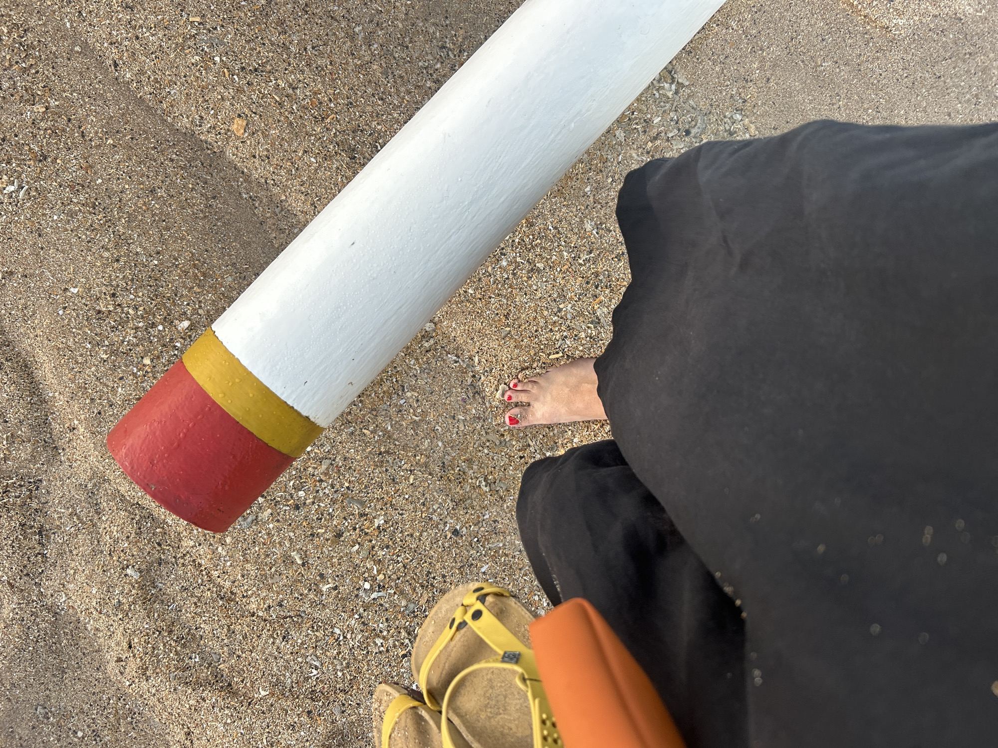 A low-angle, medium shot captures a person's bare left foot with red-painted toenails standing on a sandy beach. The foot is positioned in the lower center of the frame. To the left of the foot, a black garment, possibly pants or a skirt, takes up the lower portion of the image. Above this garment and to the left of the foot, a pair of yellow sandals rests on the sand. An orange clutch bag is also visible, partially overlapping the black garment. In the upper right quadrant of the image, a thick, white cylindrical object with a red and yellow band at its closest end lies horizontally on the sand. The sand itself appears coarse, containing small shells and pebbles, and is illuminated by natural light, casting a shadow from the cylindrical object. The scene is set on a beach, likely in Denpasar, Indonesia, given the context.