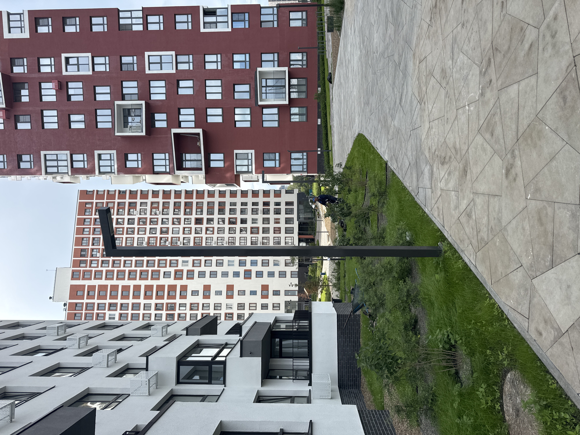 This is a ground-level, eye-level, vertical, wide-angle shot of a modern residential complex in Yekaterinburg, Russia, under an overcast sky. In the foreground, a curved, geometric-patterned concrete walkway leads towards the viewer. To the left of the walkway is a strip of lush green grass with some sparse young trees and bushes. Beyond the greenery, a white building with dark grey accents and numerous windows occupies the left side of the frame. Its balconies are enclosed with dark railings.
Further into the background, two taller apartment buildings stand side-by-side. The building on the left is predominantly white with repeating patterns of reddish-brown and dark grey rectangular windows. The building on the right is a striking deep red with white accents around many of its windows and balconies, some of which feature white open frames. The windows are uniformly rectangular and dark-tinted.
In the middle ground, between the walkway and the buildings, a person wearing dark clothing and a hat is walking while appearing to work with a tool in the grassy area. A tall, dark grey lamp post stands prominently in the center of the frame, obscuring some of the view of the buildings behind it. The overall scene suggests a newly developed urban residential area with a focus on modern architecture and landscaping.