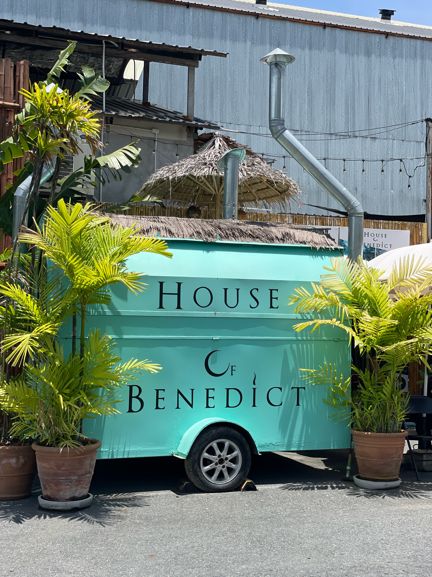 An eye-level, medium shot captures a teal-colored food truck with the words "HOUSE" and "BENEDICT" emblazoned in black serif font, with a stylized "C" and "F" in between. The truck is parked on an asphalt surface in Pattaya City, Thailand, under a bright, sunny sky. To the left and right of the truck are large potted plants with lush green fronds, their shadows cast onto the ground. Behind the food truck, a rustic thatched roof structure with a metal vent pipe rising from it is visible. The background consists of a corrugated metal building, some bamboo fencing, and string lights overhead. The scene appears to be an outdoor dining or food vendor area.