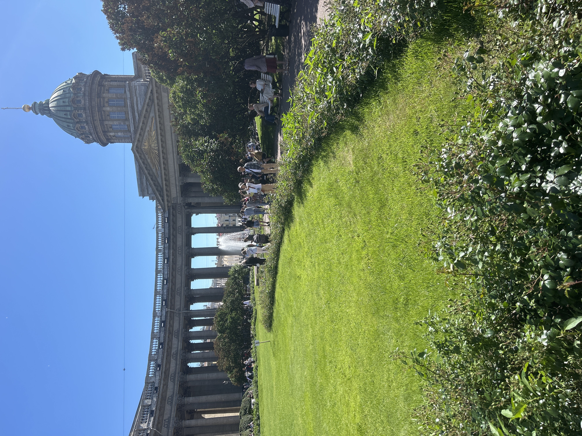 In a sunlit outdoor scene, a large green lawn is situated in front of a grand, semicircular colonnade of classical columns. The colonnade is topped with a balustrade and a curved upper story. To the right of the colonnade, a large green dome with a golden spire rises above a neoclassical building, characteristic of St. Petersburg's architectural style. In the background, a fountain sprays water into the air, and a crowd of people are visible, some walking and others sitting on benches in the shade of trees. The foreground is occupied by dense green bushes and a well-maintained lawn. The overall impression is of a vibrant public space on a clear, sunny day, likely a popular tourist destination.