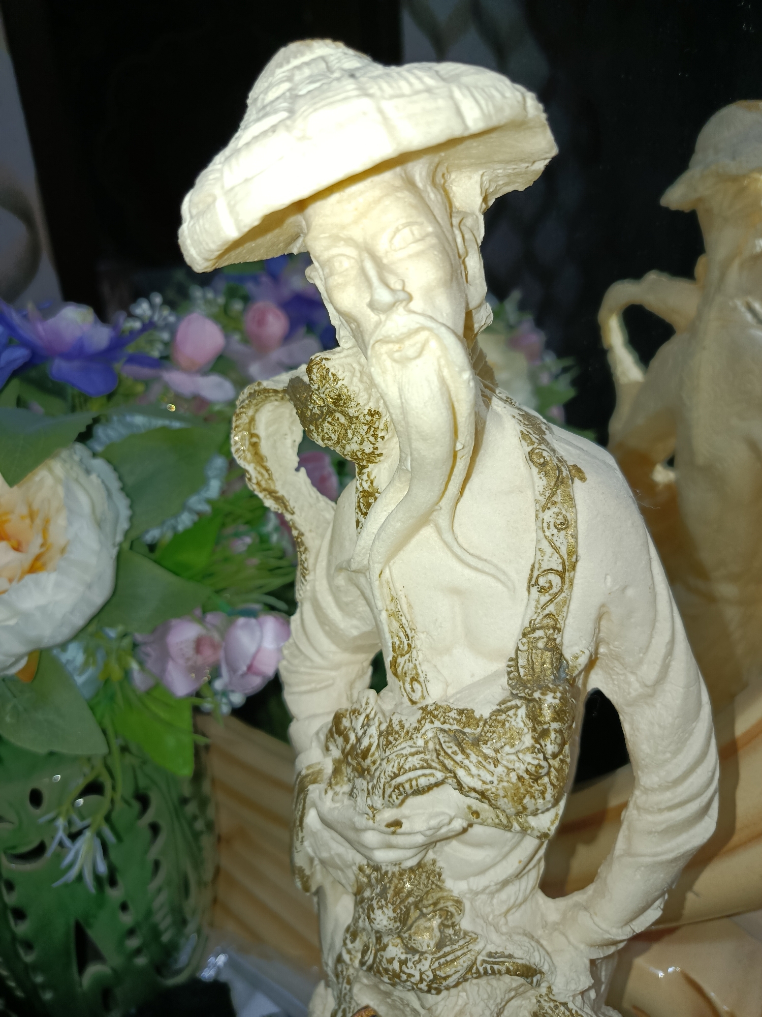 The image displays a detailed, cream-colored statuette of an elderly Asian man wearing a conical hat, a long beard, and ornate clothing. The statuette is positioned in the foreground, with its right side facing the viewer. The man's left hand is clasped at his waist, and his right arm is bent, with his hand resting on his hip. He appears to be dressed in traditional robes adorned with intricate golden patterns, depicting what looks like a dragon or serpent motif across his chest and shoulders. The statuette is slightly aged, with some areas showing a weathered texture and hints of gold paint.
Behind the statuette, to the left, is a vibrant arrangement of artificial flowers, featuring large white and yellow roses, and smaller pink and purple blossoms, interspersed with green foliage. To the right of the statuette, another similar cream-colored statuette is partially visible, suggesting they might be part of a collection. The background is dark and indistinct, with hints of reflective surfaces, possibly glass or a mirror. The overall scene is dimly lit, with a warm light source illuminating the statuette from the right. The image does not provide any information about its location within Tashkent, Uzbekistan.
