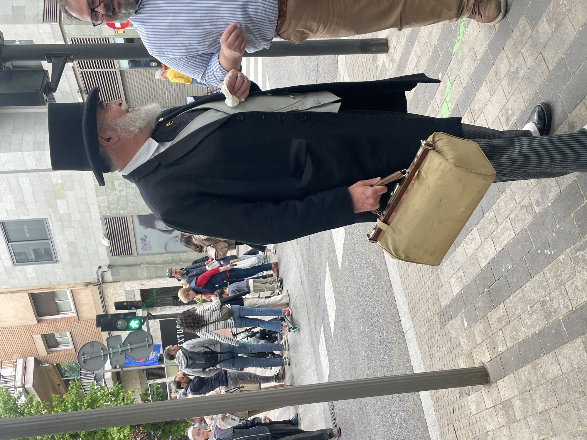 The image captures a street scene in Terrassa, Spain. A man dressed in historical attire, including a top hat, long black coat, and carrying a vintage-style bag, stands on a crosswalk. He is conversing with another man in a blue striped shirt. Behind them, a group of people are waiting to cross the street, indicating they are at a traffic light, which is currently green. The surrounding environment includes buildings with brick facades and commercial storefronts. The pavement is a patterned mix of gray and white bricks.
