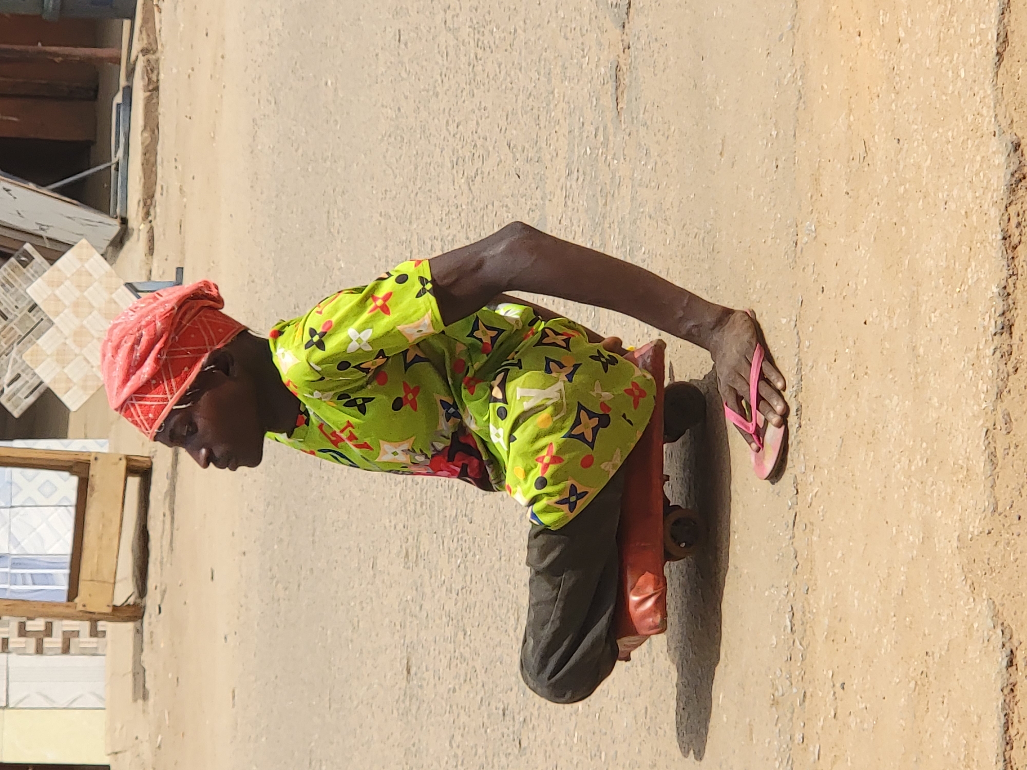 A person kneels on what appears to be a handmade skateboard on an unpaved surface next to a building. The individual wears a red head covering, a green patterned shirt, and dark pants. One foot is clad in a pink flip-flop, positioned on the ground to potentially propel the makeshift skateboard. The location seems to be outdoors, with a building's exterior visible to the left. The ground surface is pale and textured, likely made of dirt or concrete. There is no direct indication of the specific city of Maiburiji, Nigeria, but the setting suggests a developing urban or rural environment.
