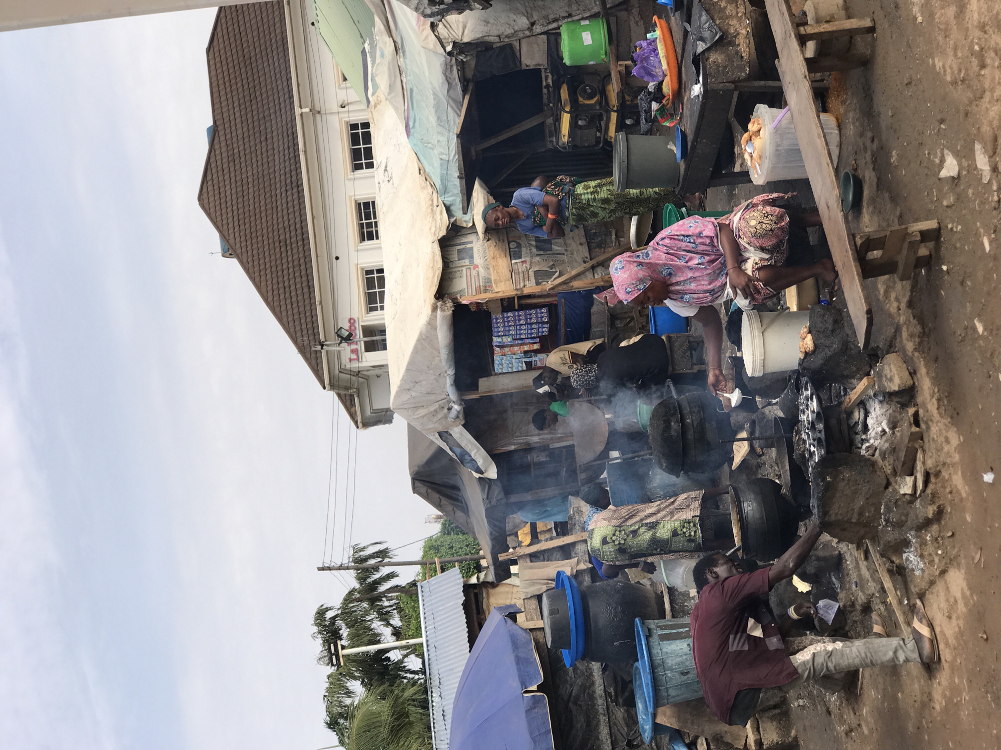The image captures a street scene in Dutsen Alhaji, Nigeria, where several people are engaged in cooking activities. At least four individuals are visible, each positioned around cooking setups consisting of large black pots placed on makeshift stoves constructed from rocks and wood.
A woman wearing a pink headscarf is near the center, tending to a pot on the fire. Another person squats near a large blue bucket, appearing to be involved in the cooking process. Smoke emanates from the cooking fires, partially obscuring the scene.
The background includes market stalls covered with tarpaulin and a building with a brown tiled roof. An assortment of items are placed on the stalls. The ground appears to be dirt. A blue umbrella is visible in the foreground. Palm trees are seen on the left.