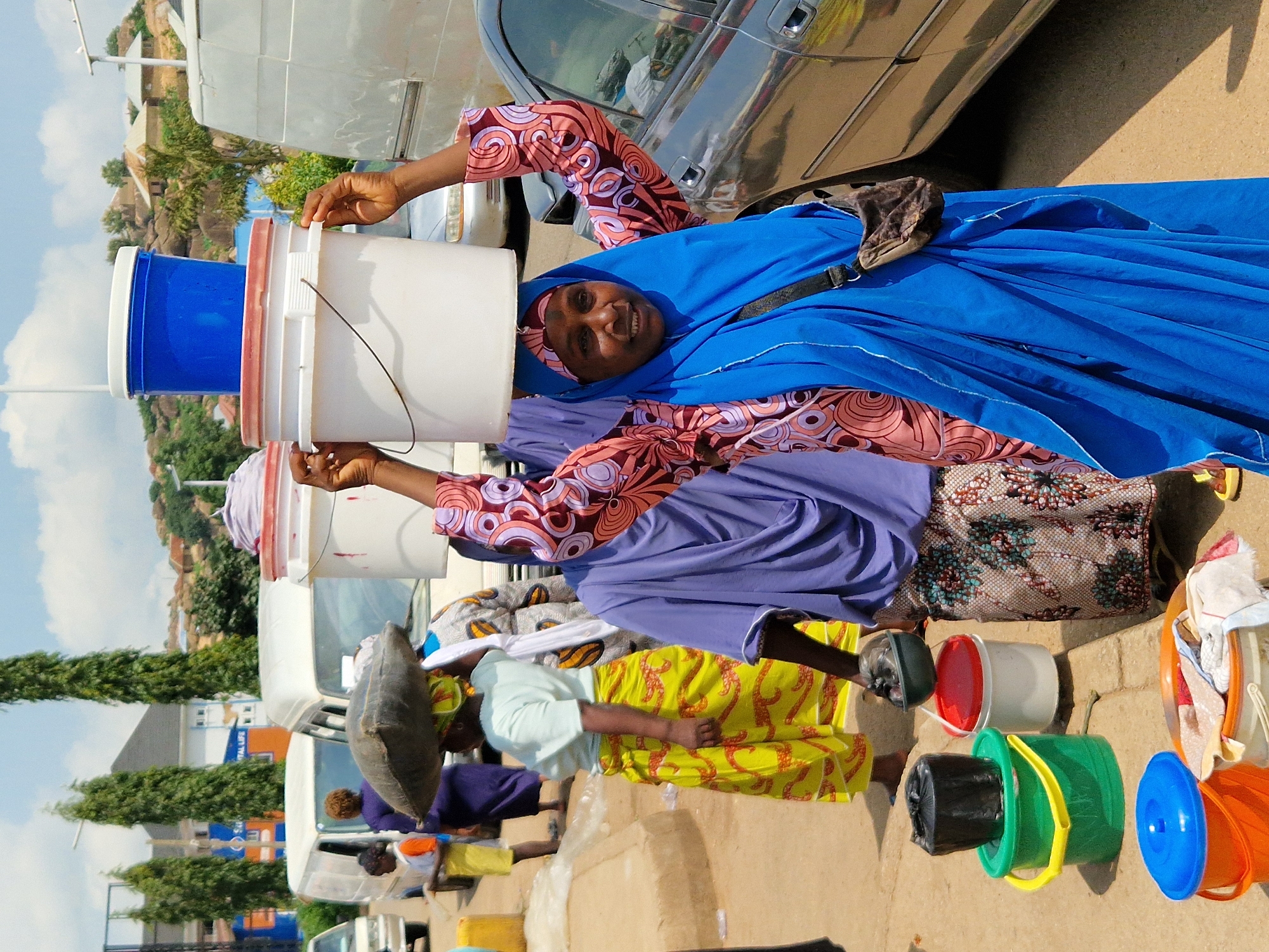 In Kabong, Nigeria, on a sunny day, a group of individuals are engaged in commerce near a street lined with parked vehicles and buildings. The foreground features several individuals, primarily women, carrying buckets on their heads. One woman, wearing a blue hijab and a vibrant pink and orange patterned dress, balances two large white buckets. Her arms are raised, supporting the load, and she looks towards the camera with a slight smile. To her left, another woman in a purple garment carries a large, dark sack on her head, her face obscured. A woman in a yellow and red patterned skirt is also visible, her head also laden with a dark object.
In the midground, a white van is parked, and a young girl in an orange and white outfit stands near it. Another white van, partially visible, is parked behind the first. Further back, more buildings and trees are seen under a bright, cloudy sky.
On the ground, near the people, are several colorful buckets. A bright green bucket with yellow handles holds a black object. Next to it is a white bucket with a red lid, and behind that, a red-lidded bucket. In the lower left corner, a blue bucket with a lid sits beside an orange bucket and a partially filled white bucket containing clothes.
The environment suggests a busy marketplace or roadside vendor area. The presence of vehicles and the variety of goods being transported indicate active commerce. The scene is characterized by the vibrant colors of the clothing and buckets, set against the backdrop of the urban Nigerian landscape.